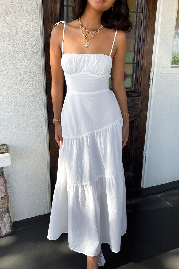 A woman wearing a white midi dress with a square neckline, tied cami straps, asymmetrical tiers, and a smocked back.
