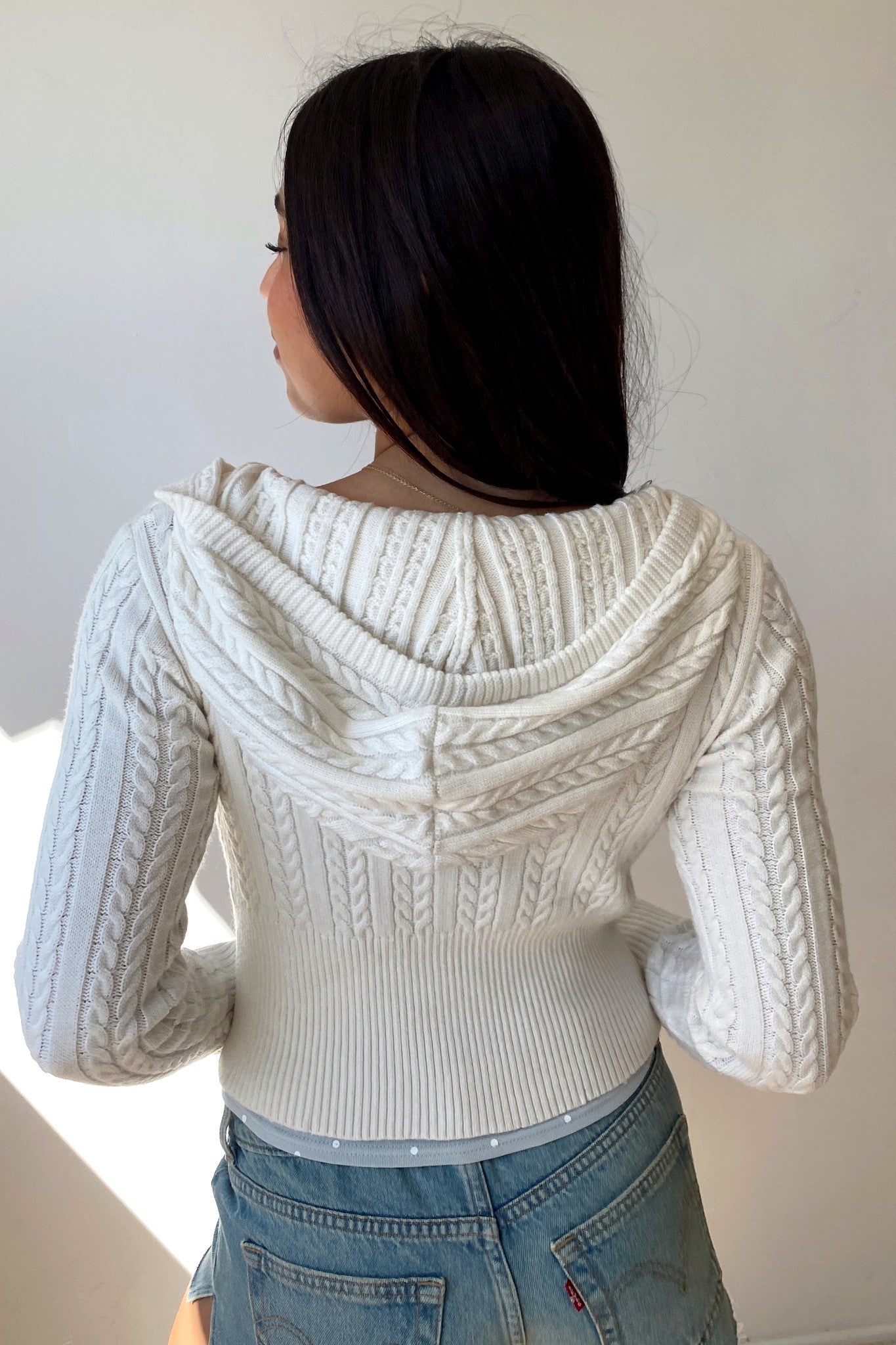 Person wearing a white cable knit sweater with a hood against a plain background
