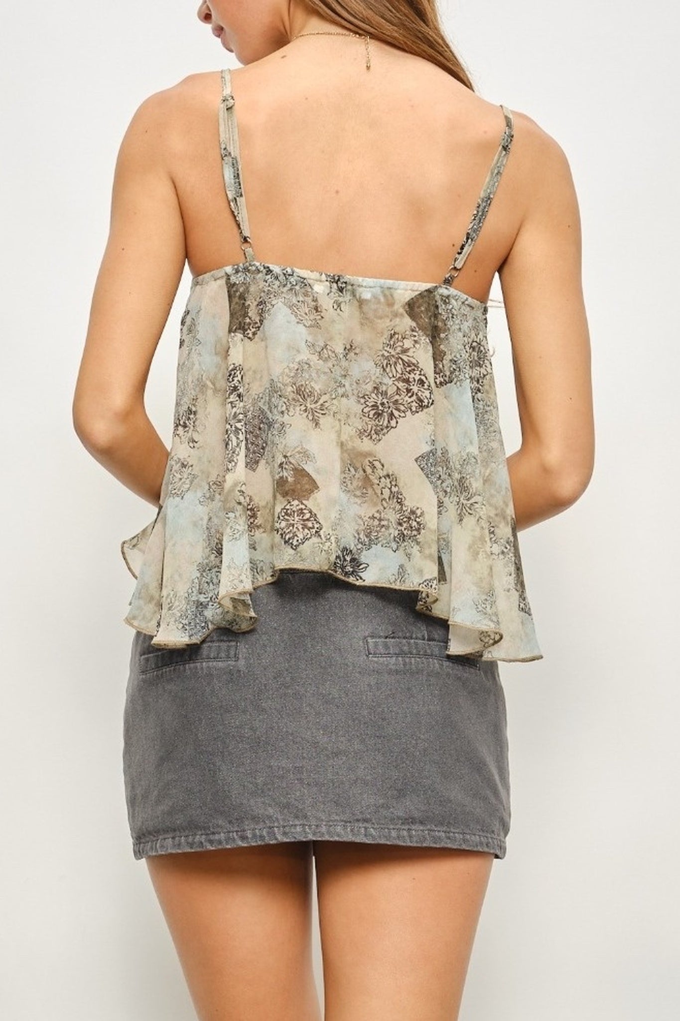 A woman shown from the back wearing a sleeveless ruffled camisole top with a beige, gray, and blue floral pattern, paired with a gray denim mini skirt, against a plain light background.