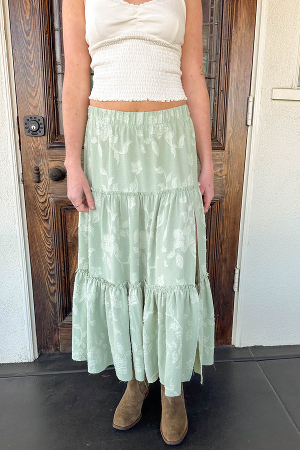 Person wearing a light green tiered skirt with a white top in front of a wooden door.