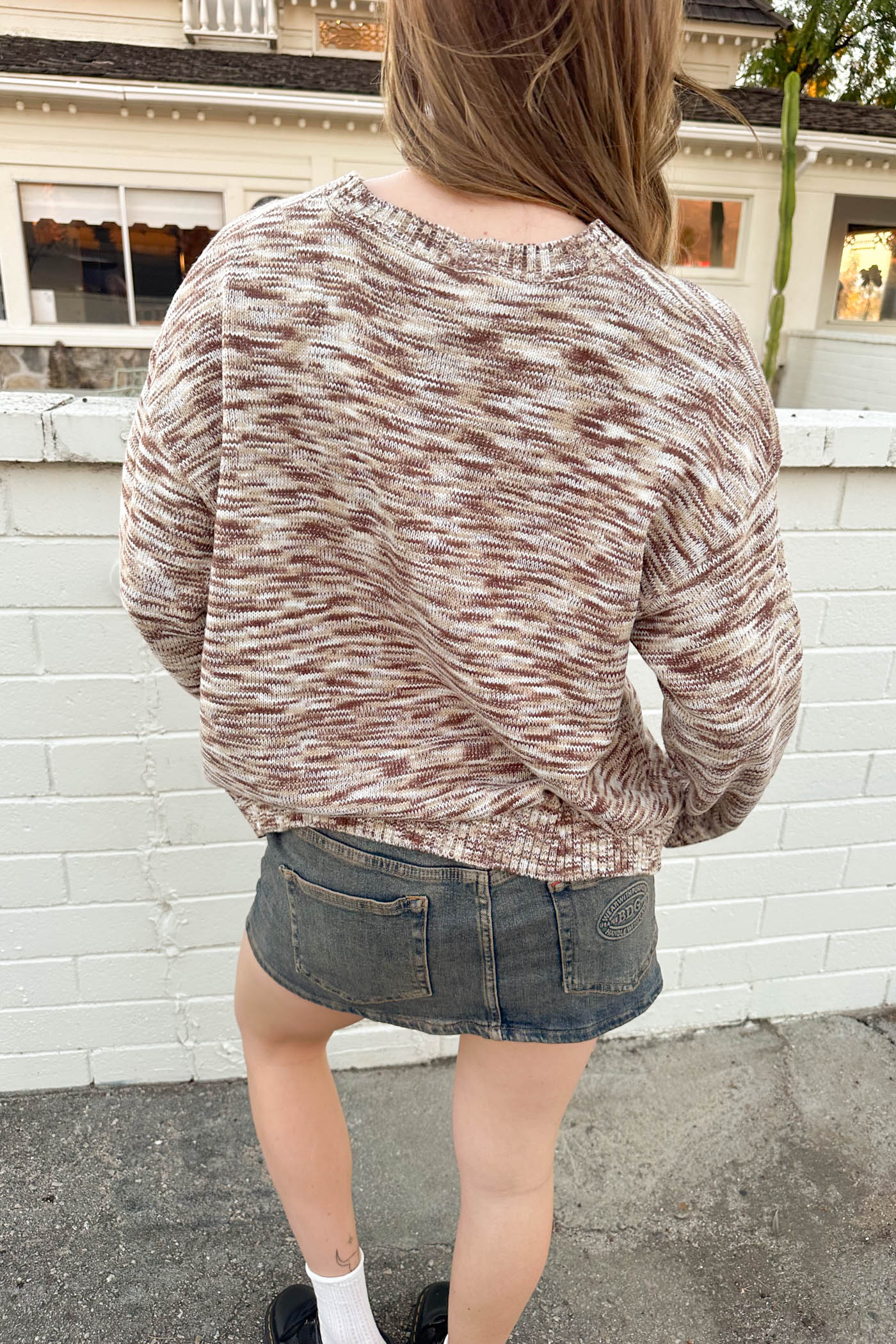 Person wearing a maroon and beige patterned sweater and denim shorts standing against a white brick wall.