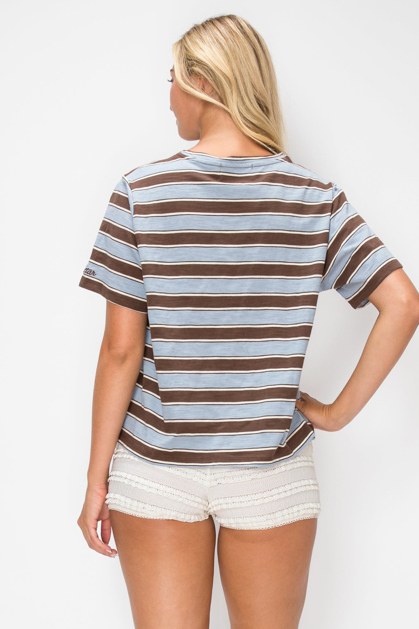 A woman shown from the back wearing a brown and light blue horizontally striped short-sleeve t-shirt and off-white shorts against a plain white studio background.