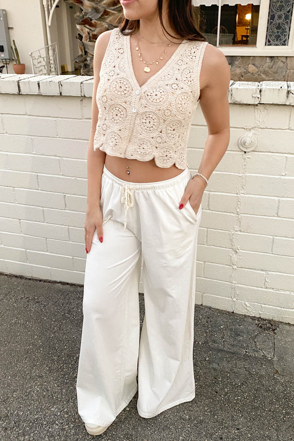 Woman wearing a beige lace top and white pants standing against a white brick wall.