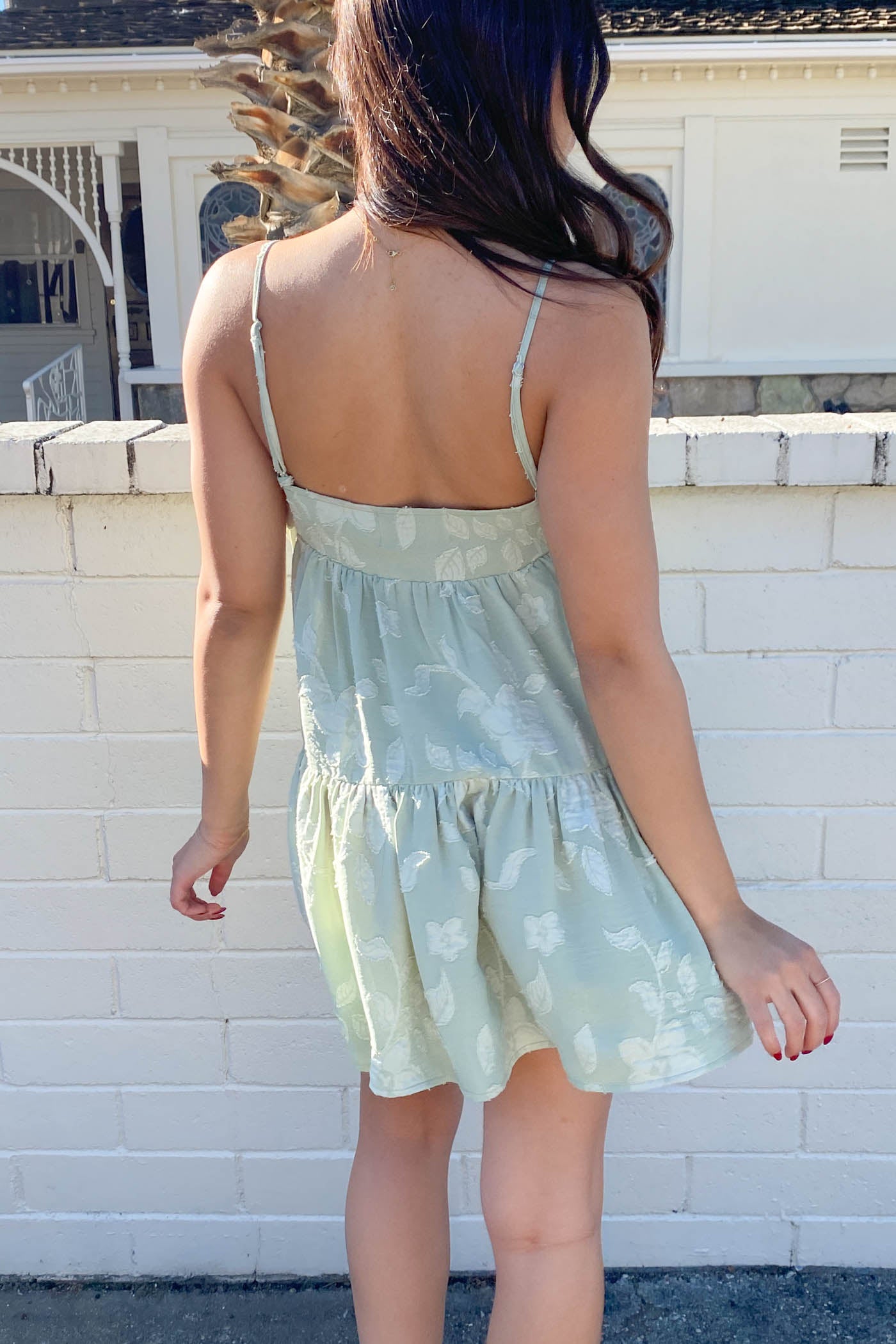 Woman wearing a light green dress with floral pattern against a white brick wall.