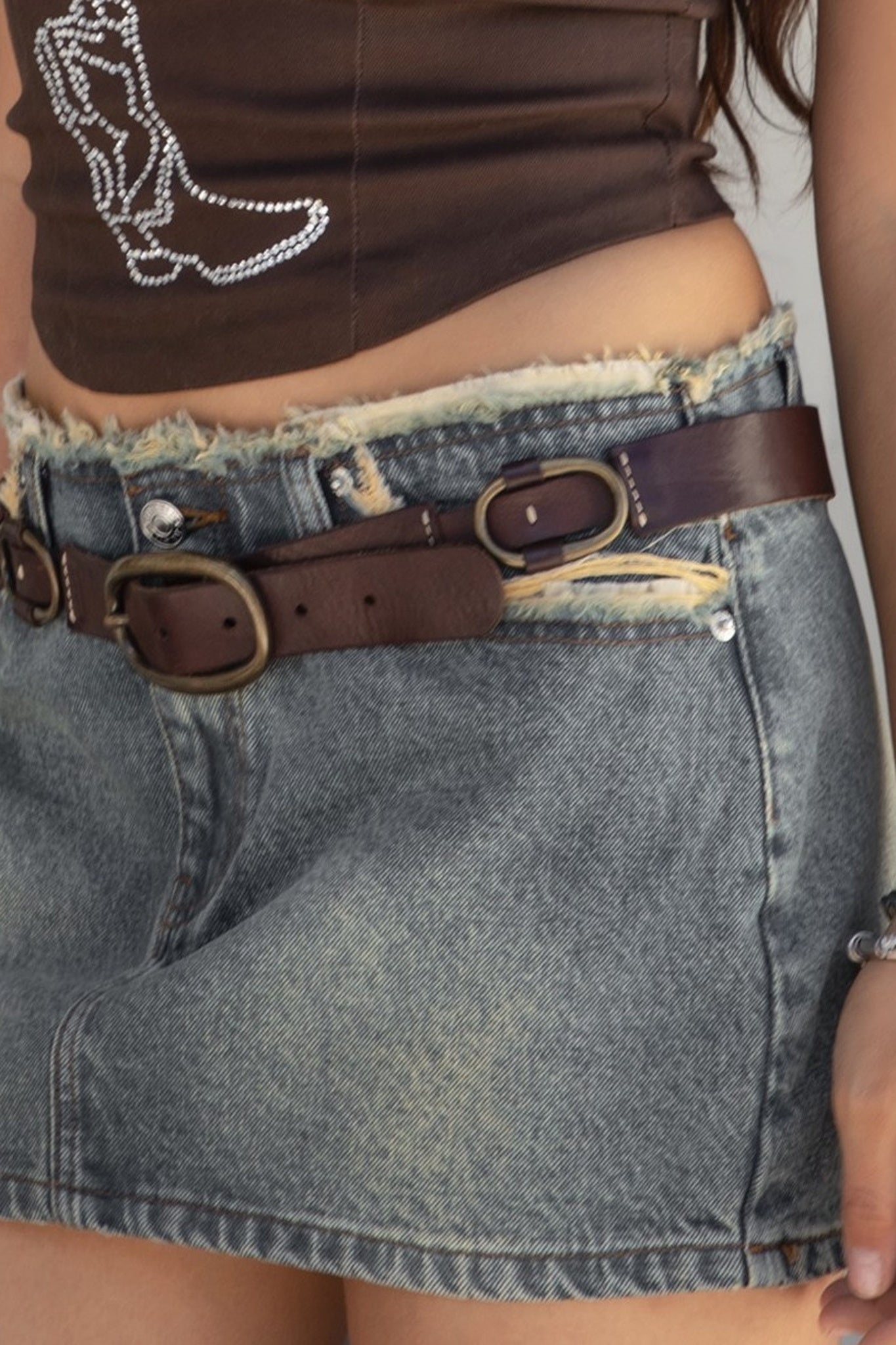 A person wearing a brown cropped tank top with 'Cowboy Cowboys' and a cowboy boot graphic, a frayed blue denim mini skirt with a brown leather belt, and brown leather cowboy boots, standing against a white wall.