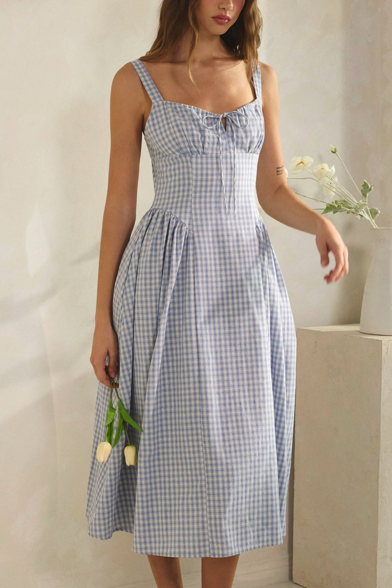 Woman wearing a blue checkered dress holding flowers against a neutral background