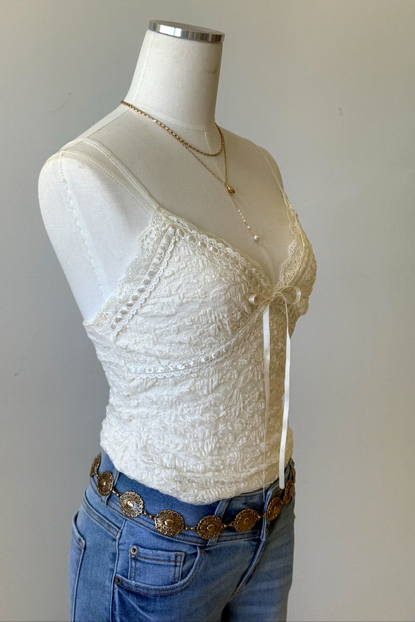 An off-white lace camisole top displayed on a white mannequin torso, styled with blue jeans and a western-style belt.