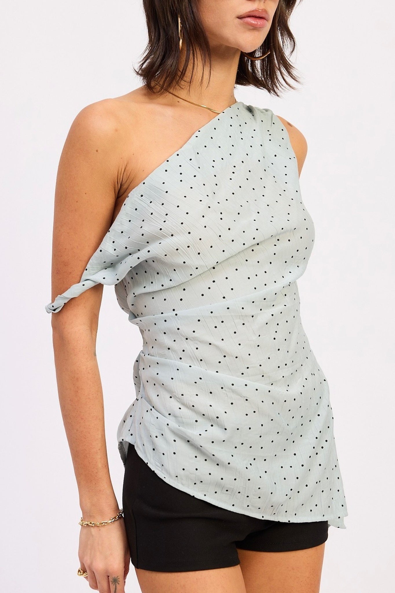 Woman wearing a light blue polka dot one-shoulder top with black shorts on a white background