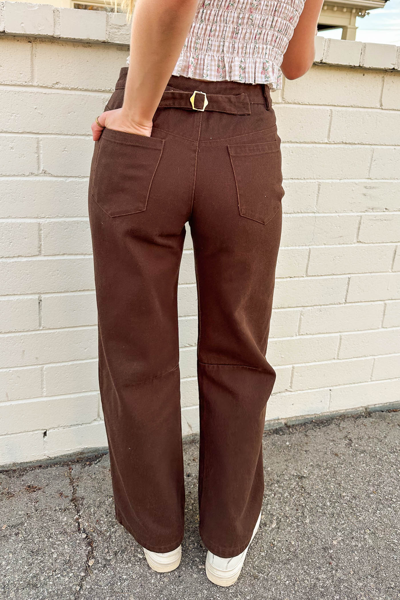 Person wearing brown pants with a belt in front of a white brick wall.