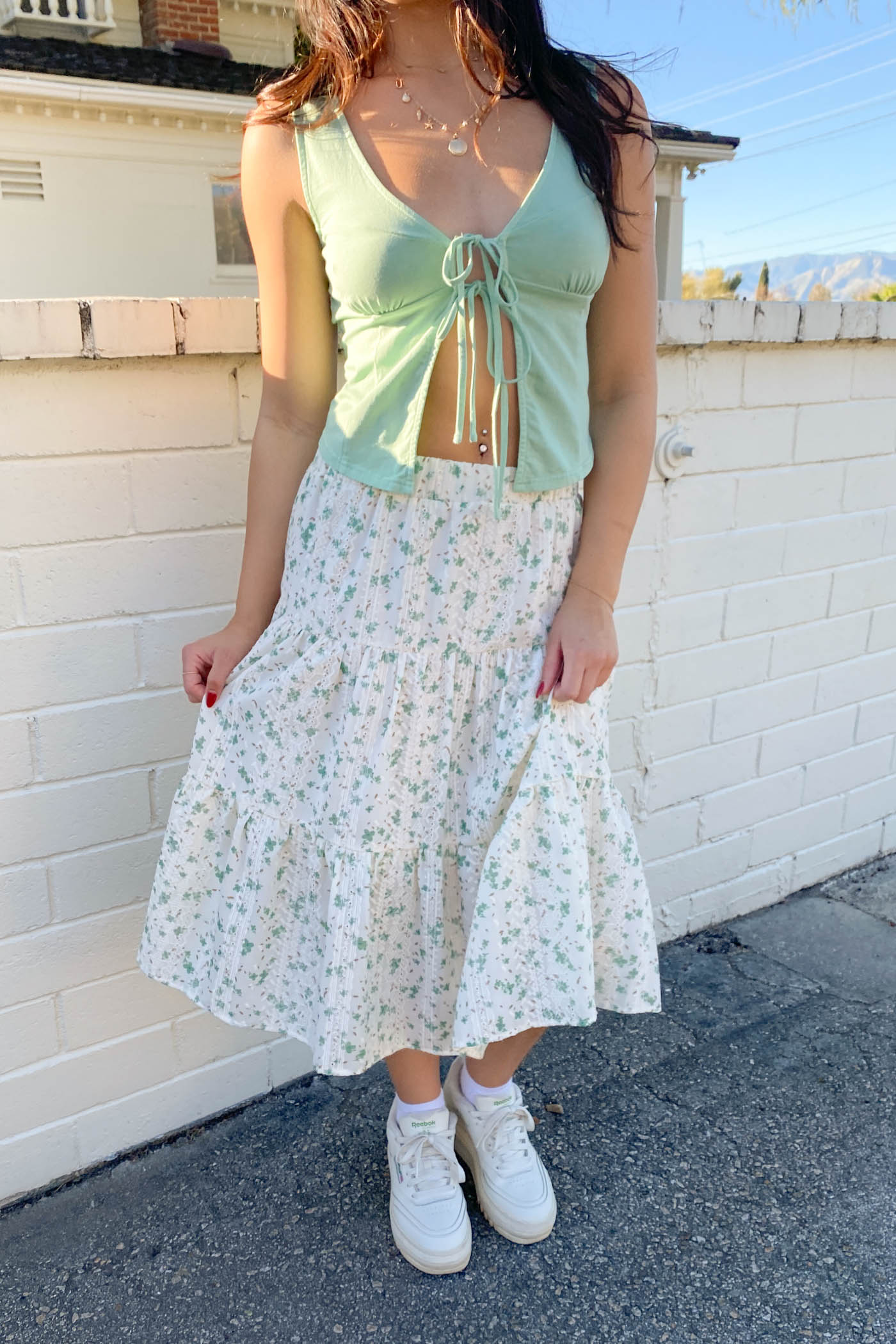 Woman wearing a light green top and floral skirt against a white brick wall.