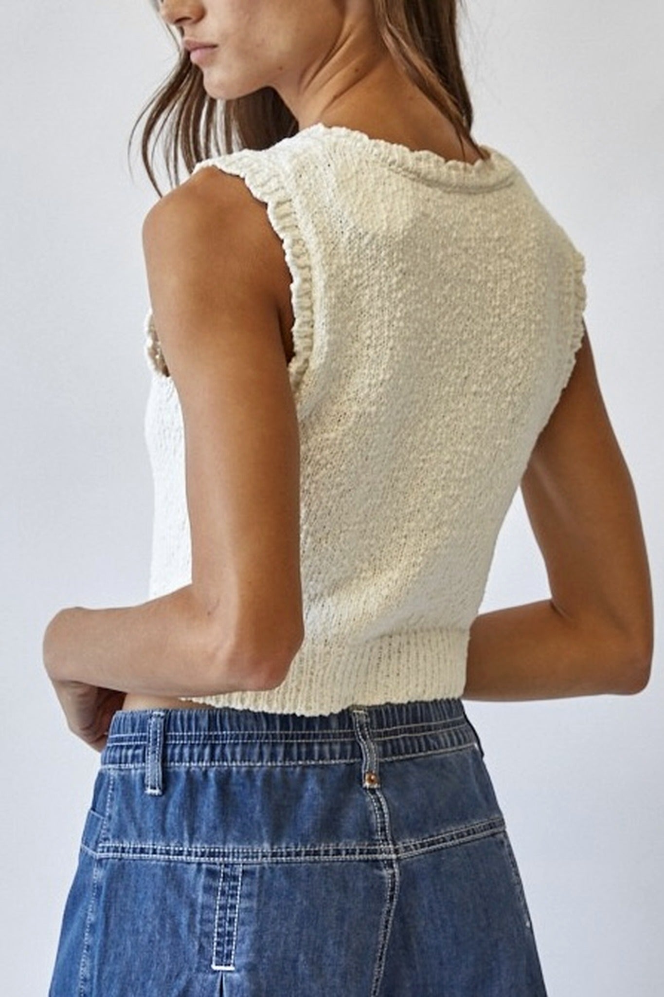 Woman wearing a cream knitted sleeveless top and blue jeans against a white background