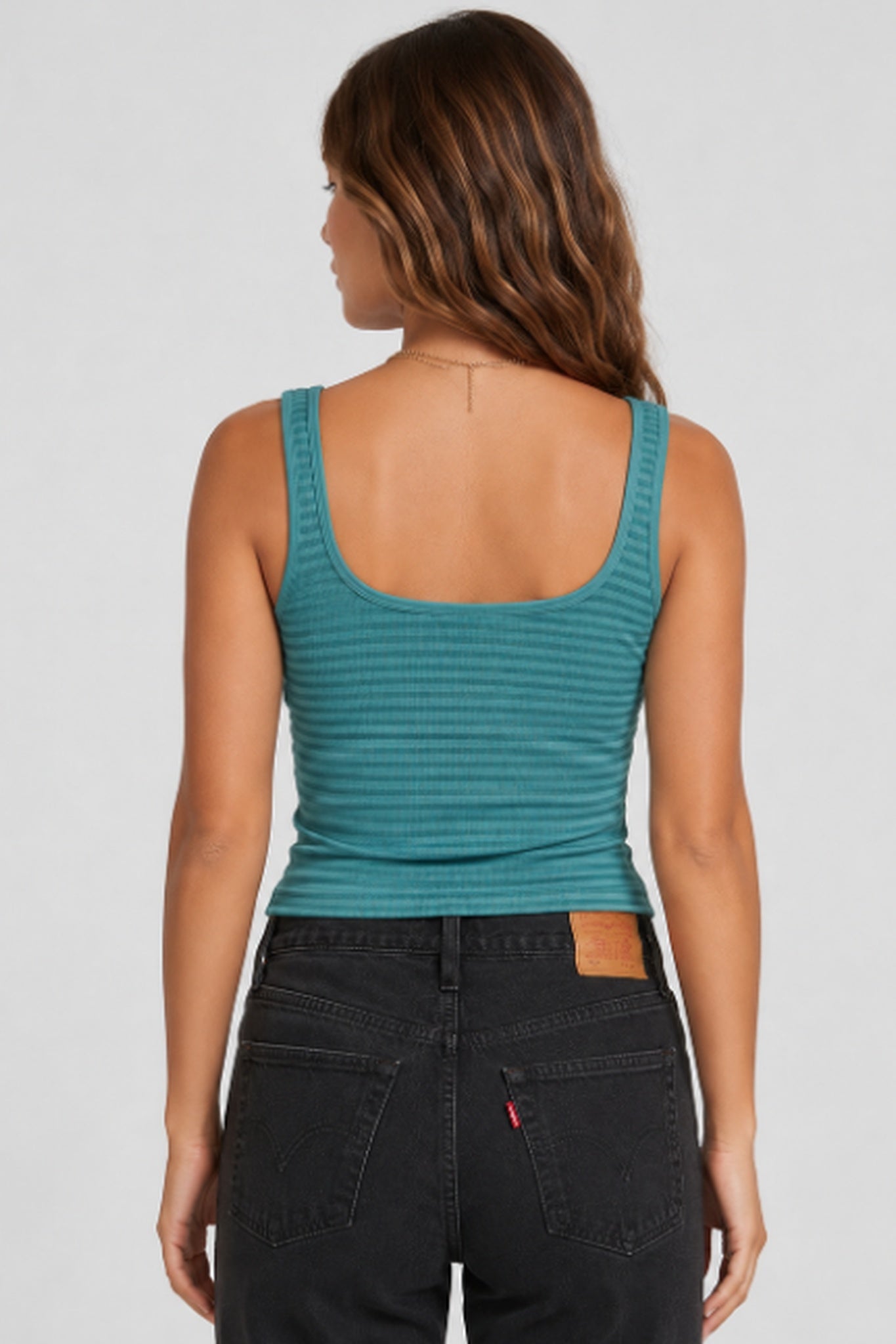 A woman shown from the back wearing a teal ribbed tank top and black jeans against a light gray studio background.