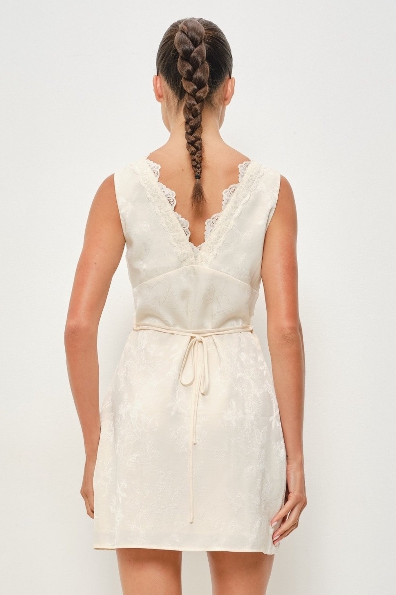 A woman shown from the back wearing a sleeveless cream-colored lace dress with a tie waist and lace-trimmed neckline, photographed against a plain light background.