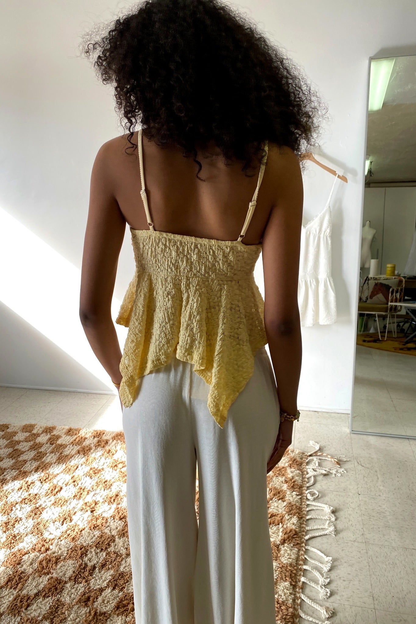 Woman wearing a yellow lace top and white pants in a room with a mirror and furniture.