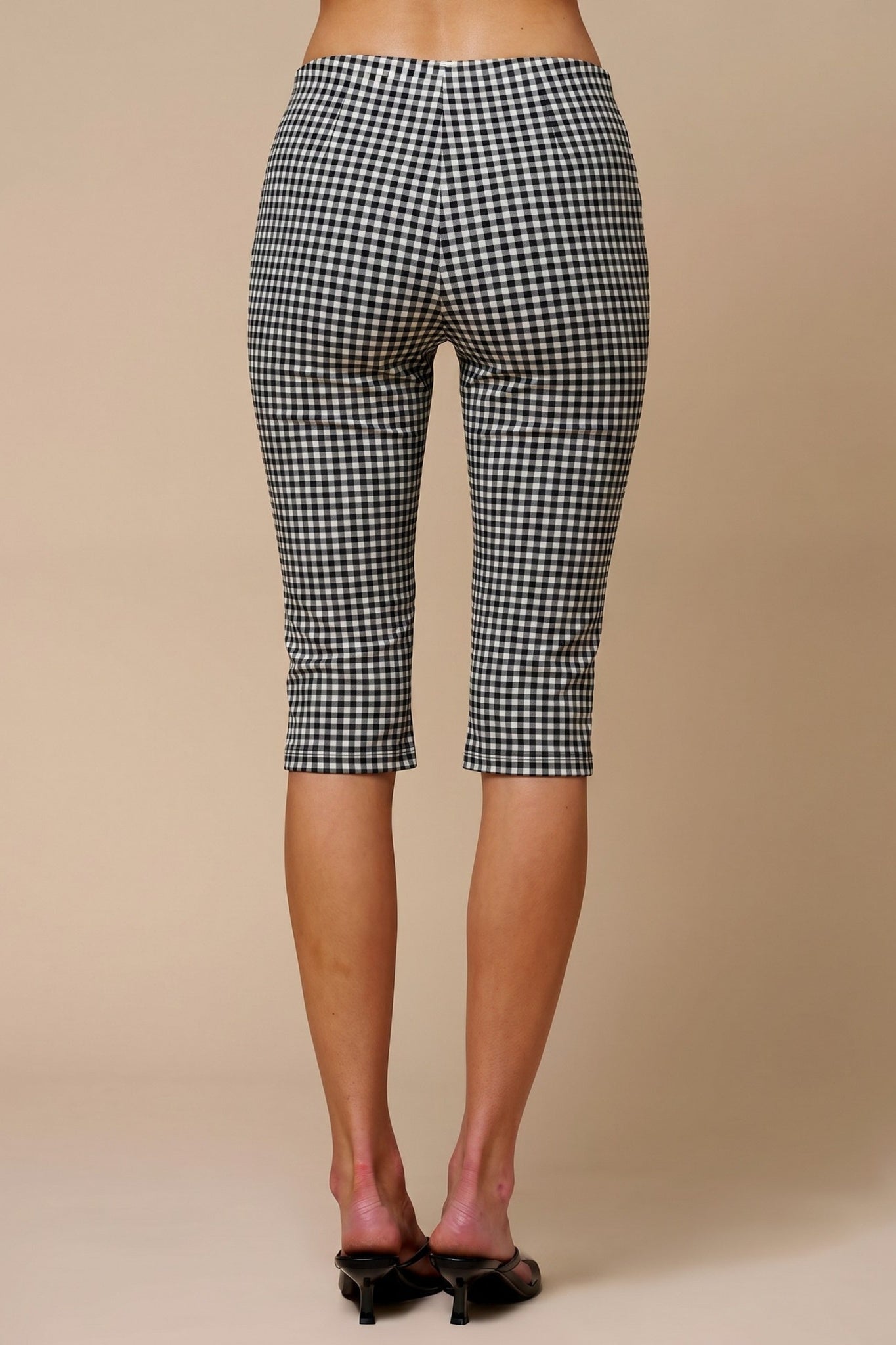 Black and white gingham capri pants shown from the back on a model, paired with black pointed-toe mule heels against a solid beige studio background.
