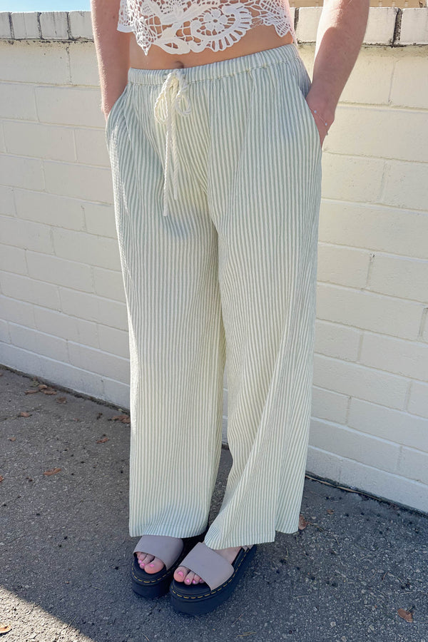 Person wearing light green striped pants and a white lace top against a white brick wall.