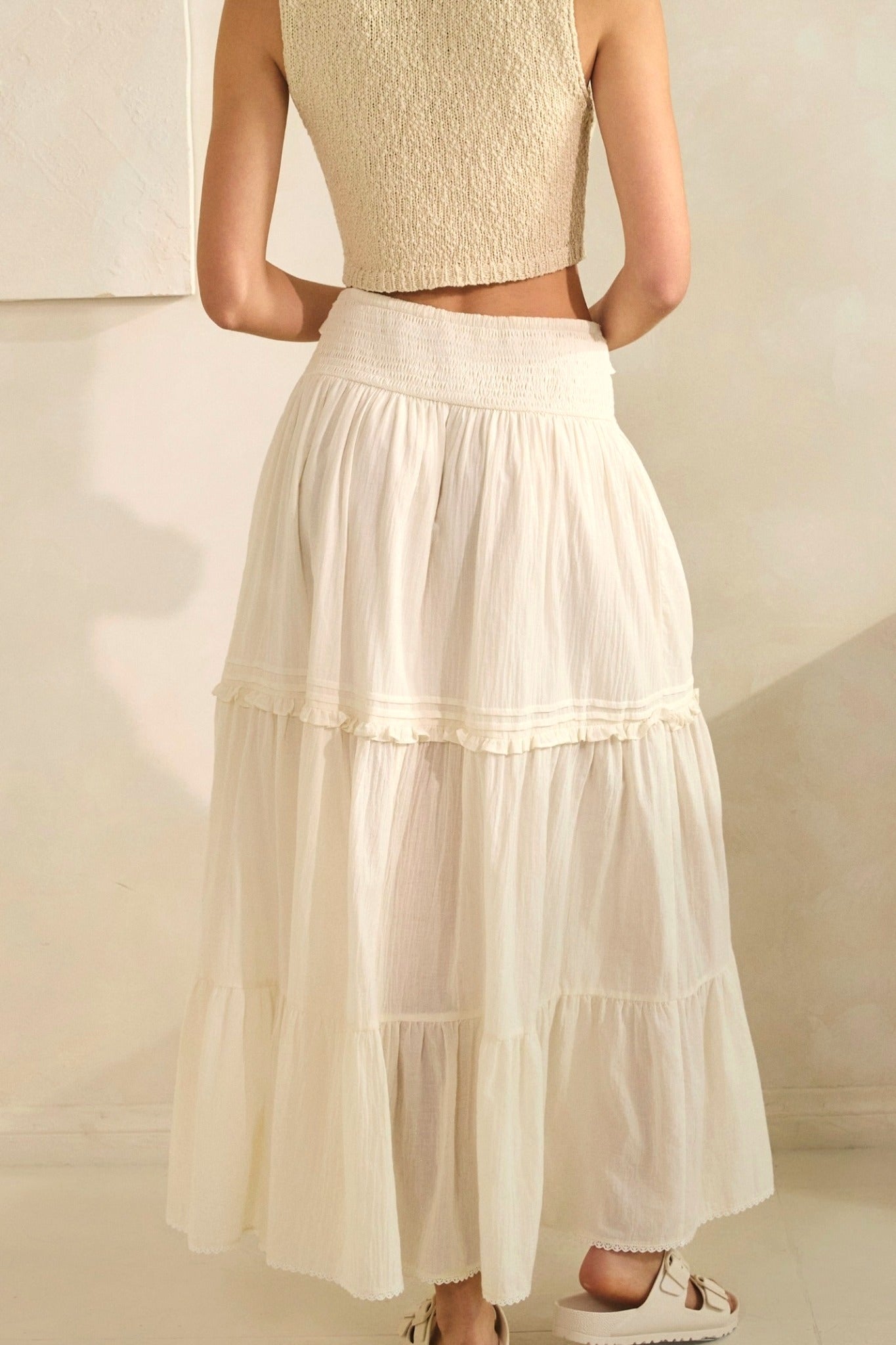 Woman wearing a beige sleeveless top and tiered cream skirt against a neutral background