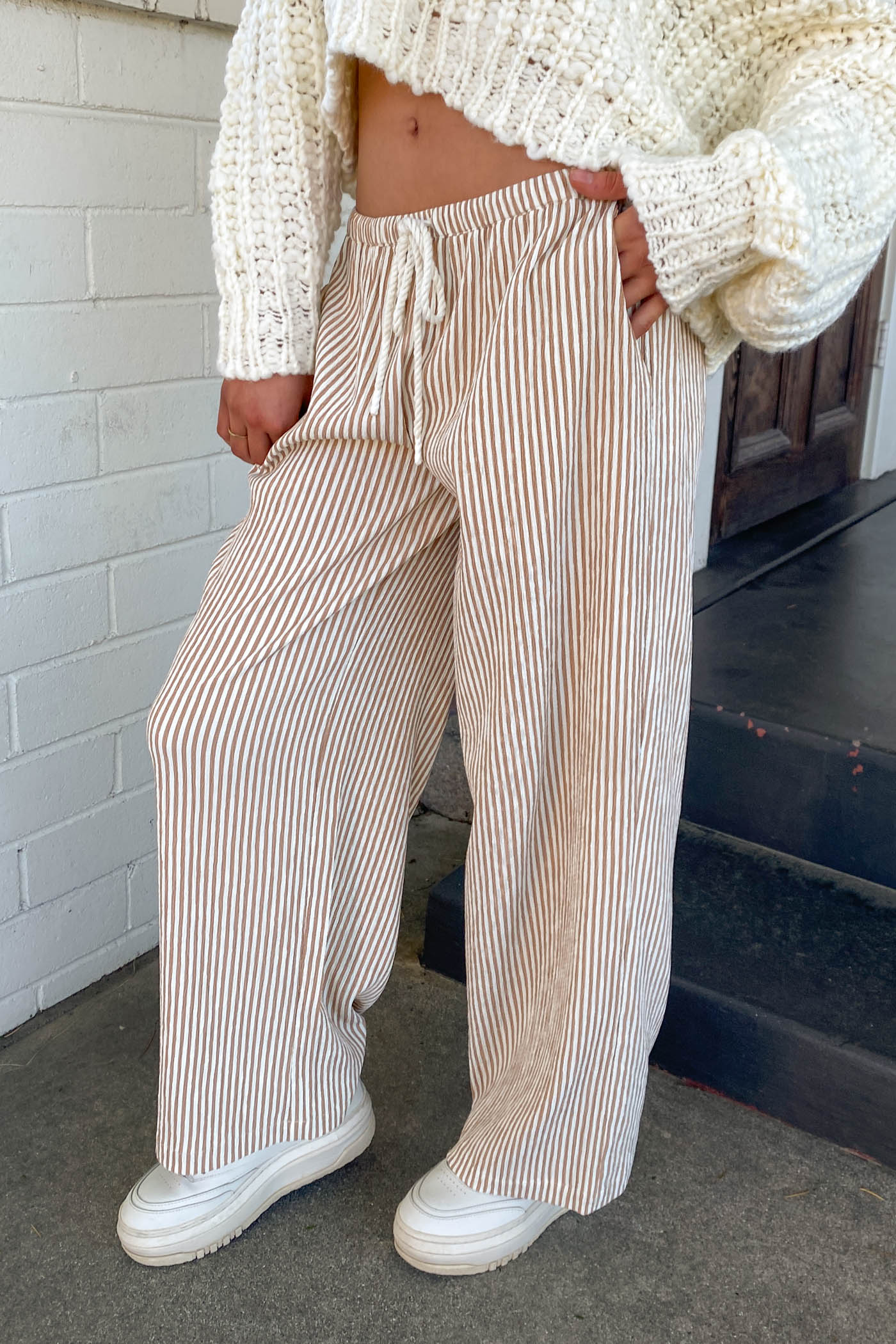 Person wearing beige striped pants and a white knit sweater standing on steps.