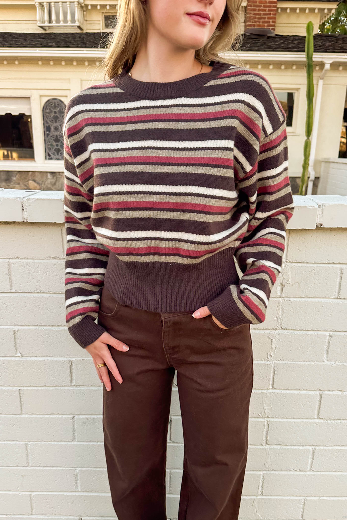 Person wearing a striped sweater and brown pants standing against a white brick wall.