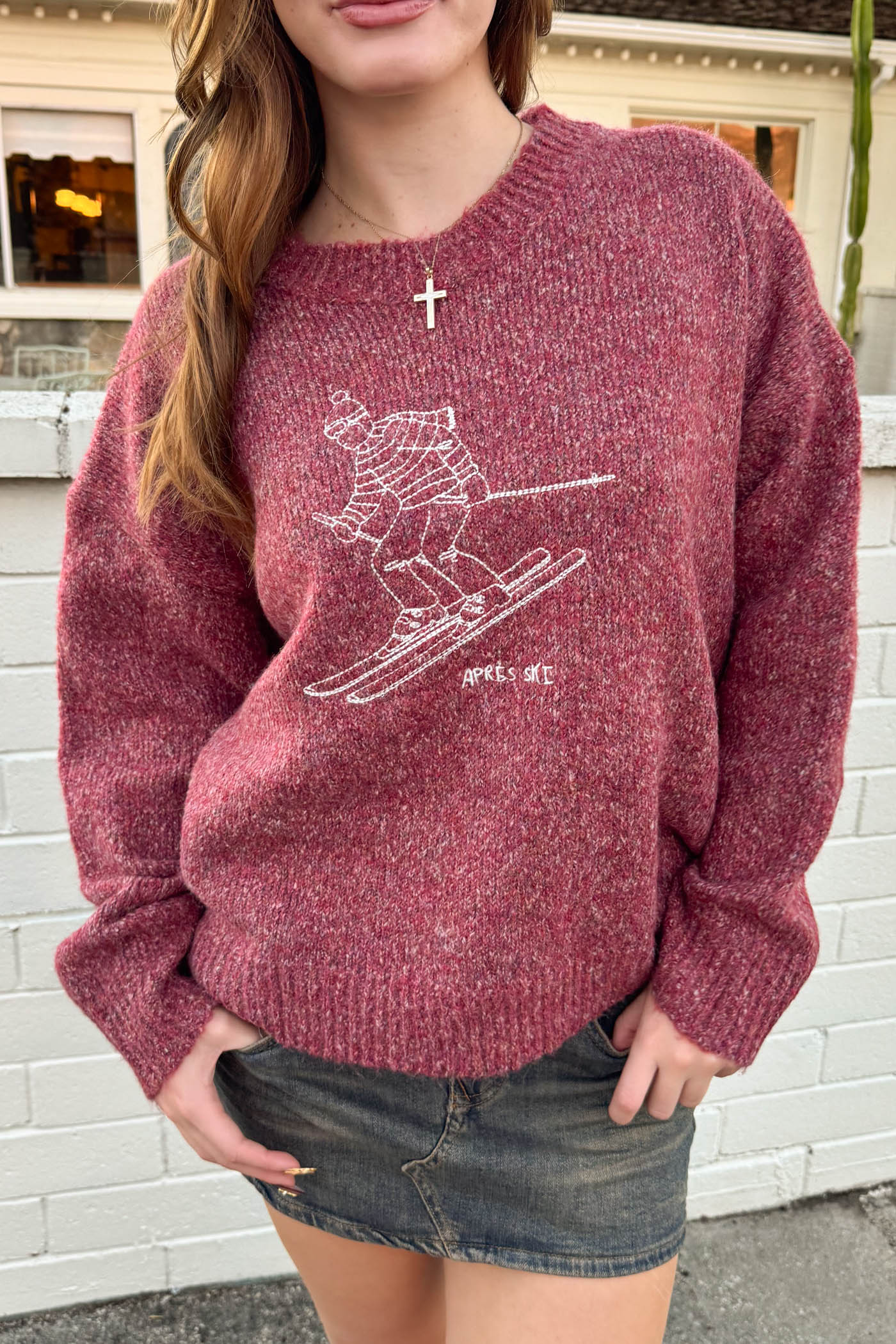 Person wearing a maroon sweater with a graphic design in front of a white brick wall.