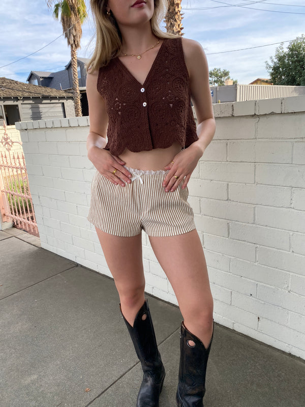 Person wearing a brown top and beige shorts with black boots on a tiled pavement.