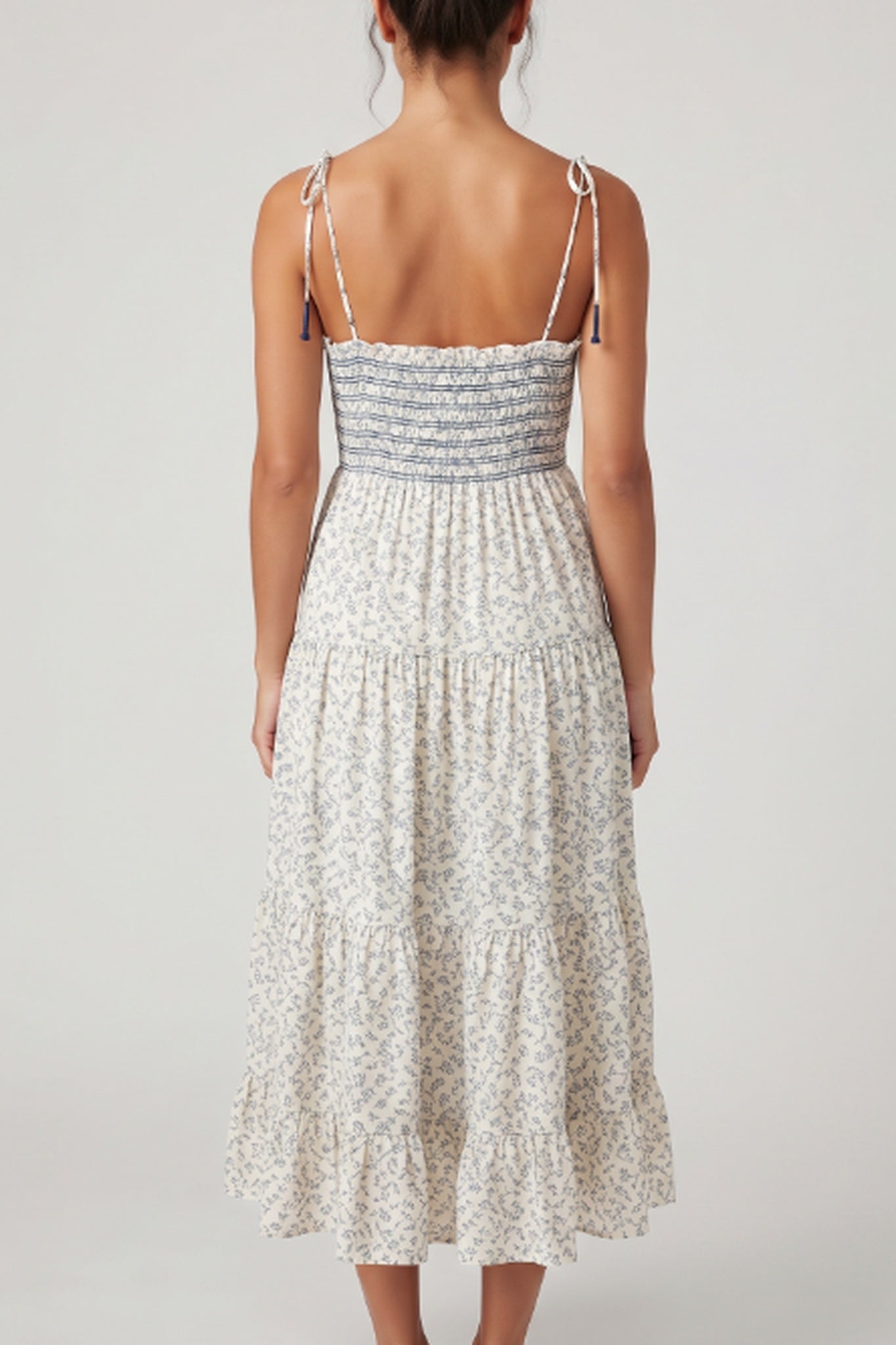 A woman shown from the back wearing a sleeveless midi dress with thin shoulder straps, a smocked bodice, and a tiered skirt in a light cream and gray floral print against a plain light gray studio background.