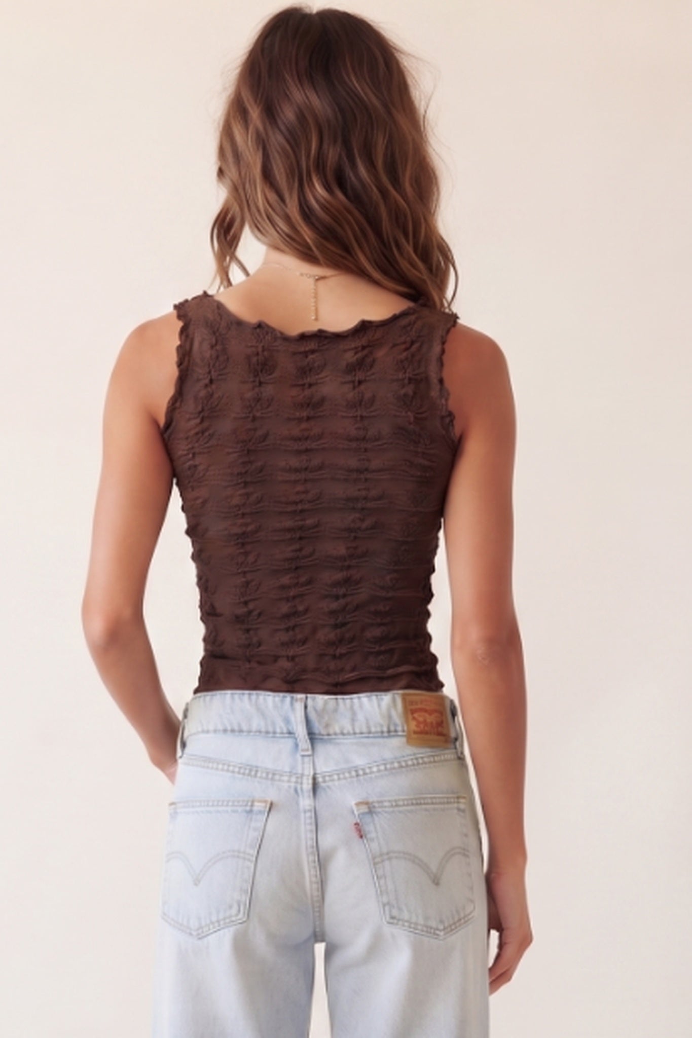 A woman shown from the back wearing a dark brown sleeveless textured top and light-wash blue jeans against a plain cream background.