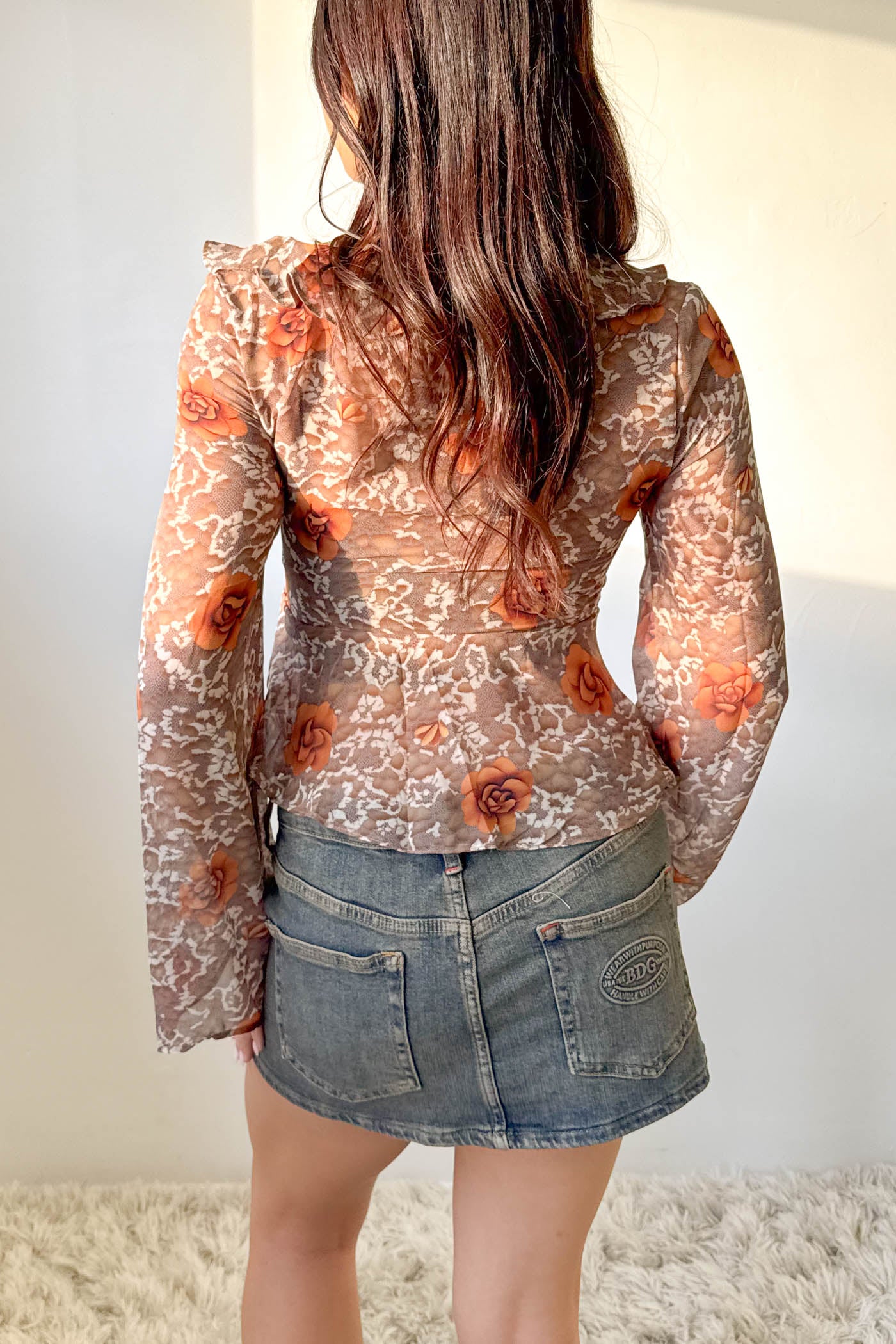 Person wearing a sheer floral blouse with long sleeves and a denim skirt against a plain background