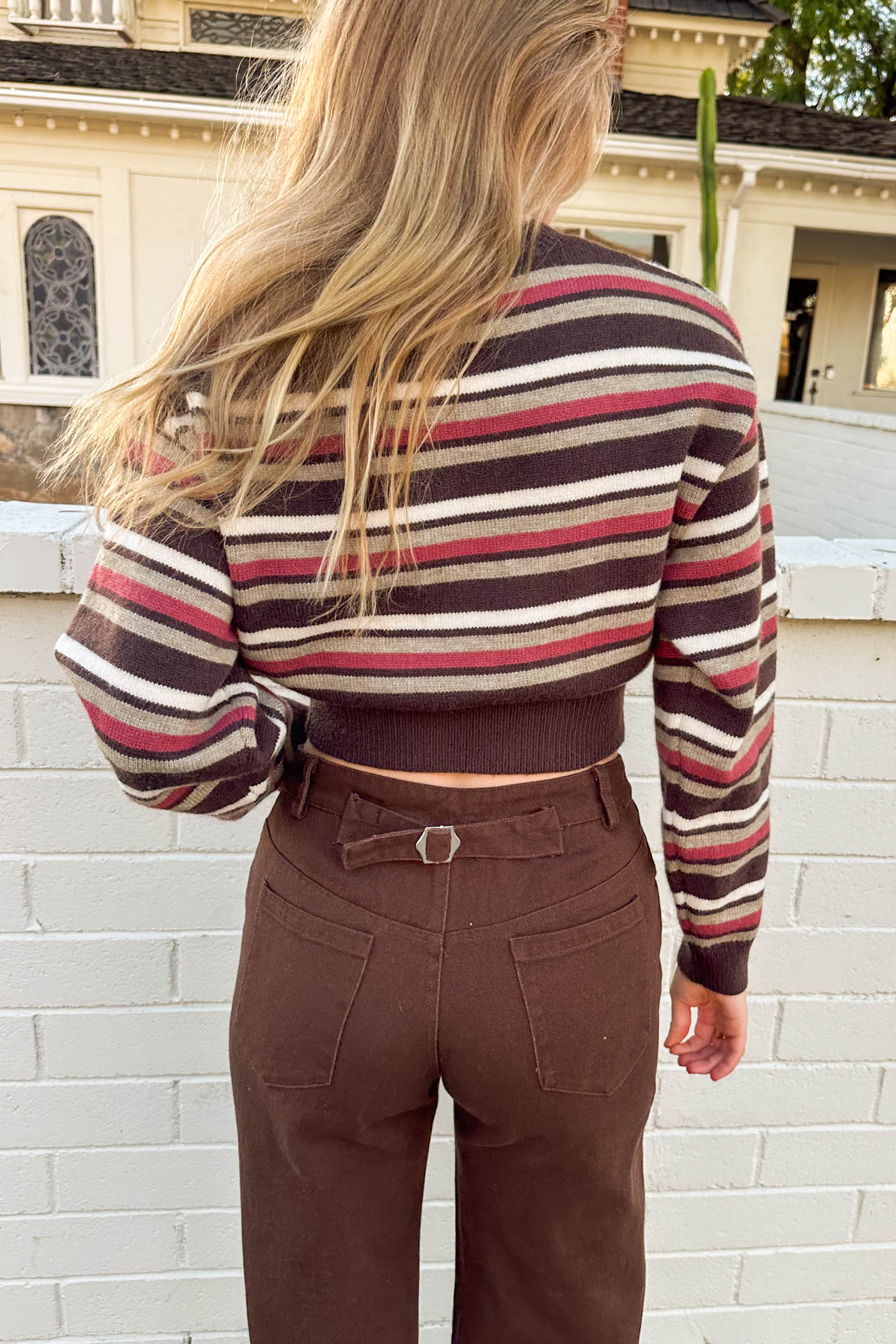 Person wearing a striped sweater and brown pants standing in front of a white brick wall.