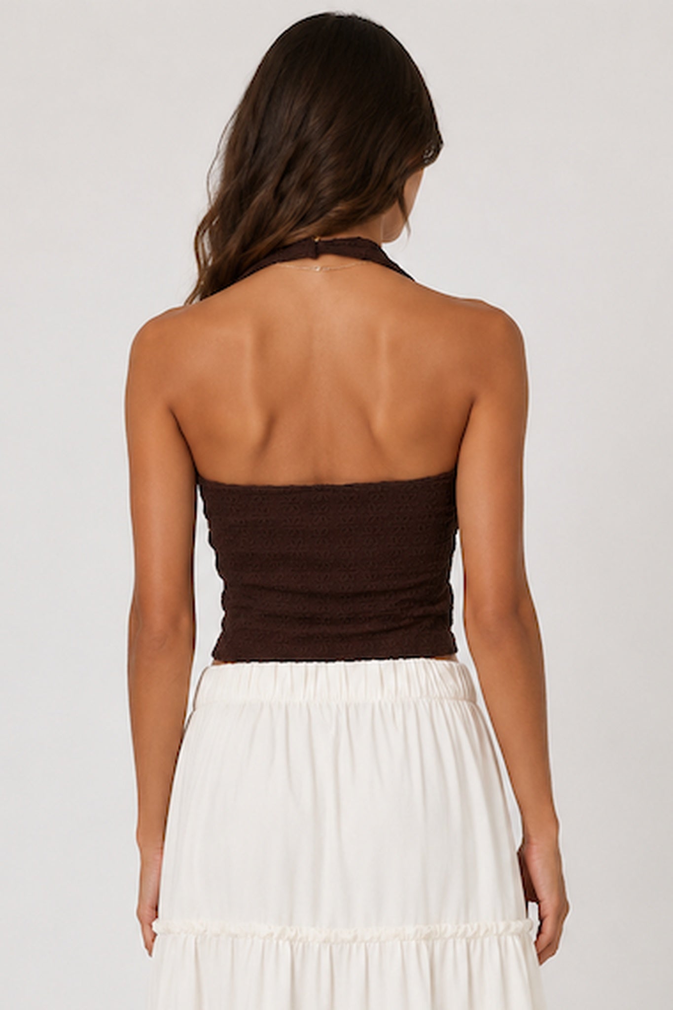 Rear view of a woman wearing a dark brown halter tube top and a white tiered skirt against a light gray studio background.
