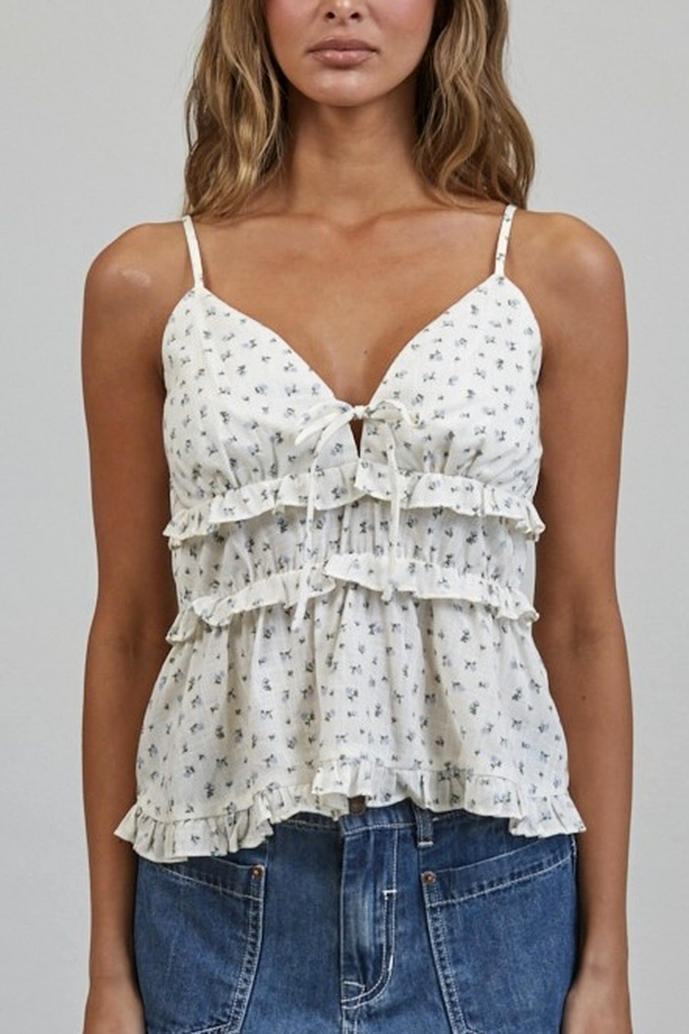 Woman wearing a white floral tiered camisole top with blue jeans on a gray background