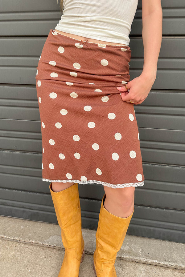 Person wearing a brown skirt with white polka dots and yellow boots against a gray background