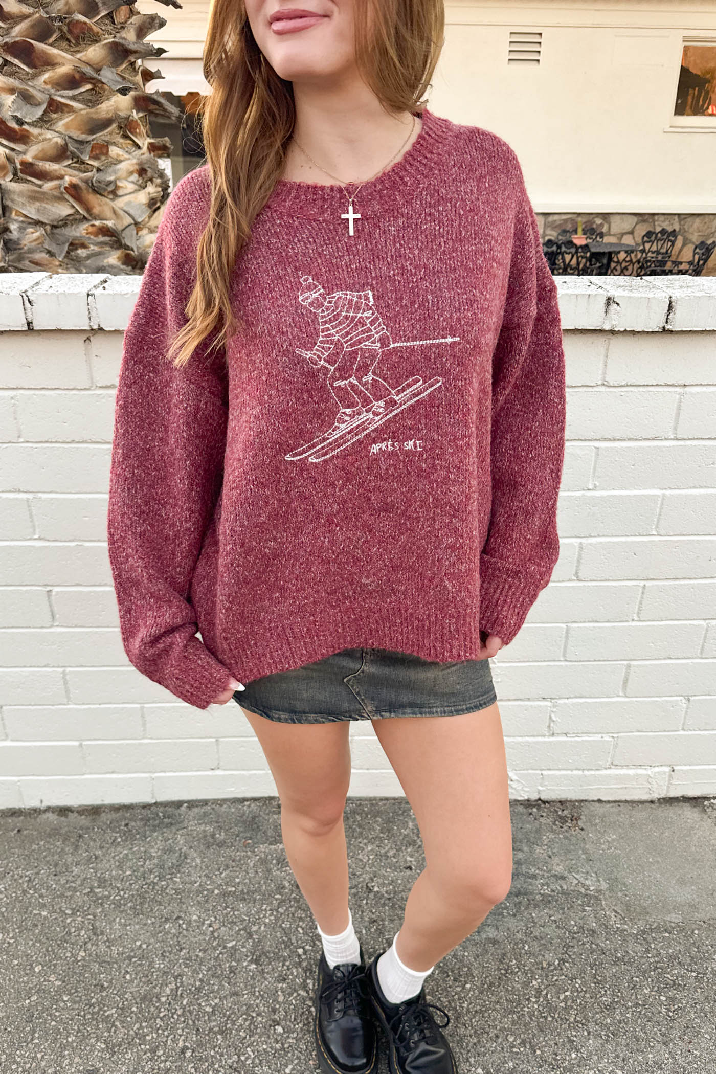 Person wearing a maroon sweater with a graphic design, standing against a white brick wall.