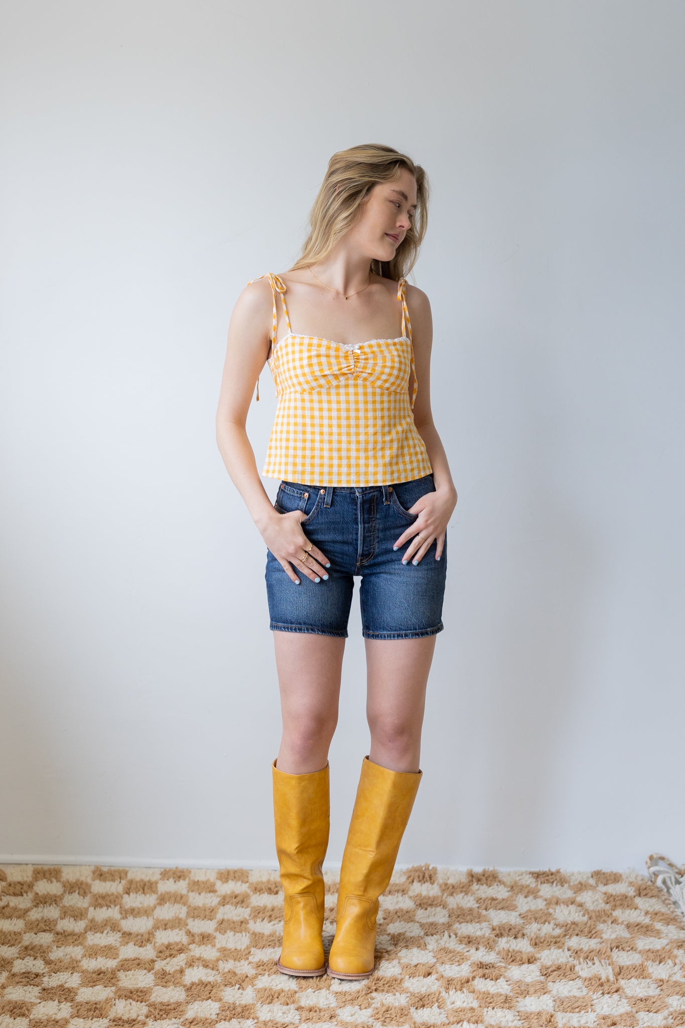 Woman wearing a yellow checkered top, denim shorts, and yellow boots against a white wall.