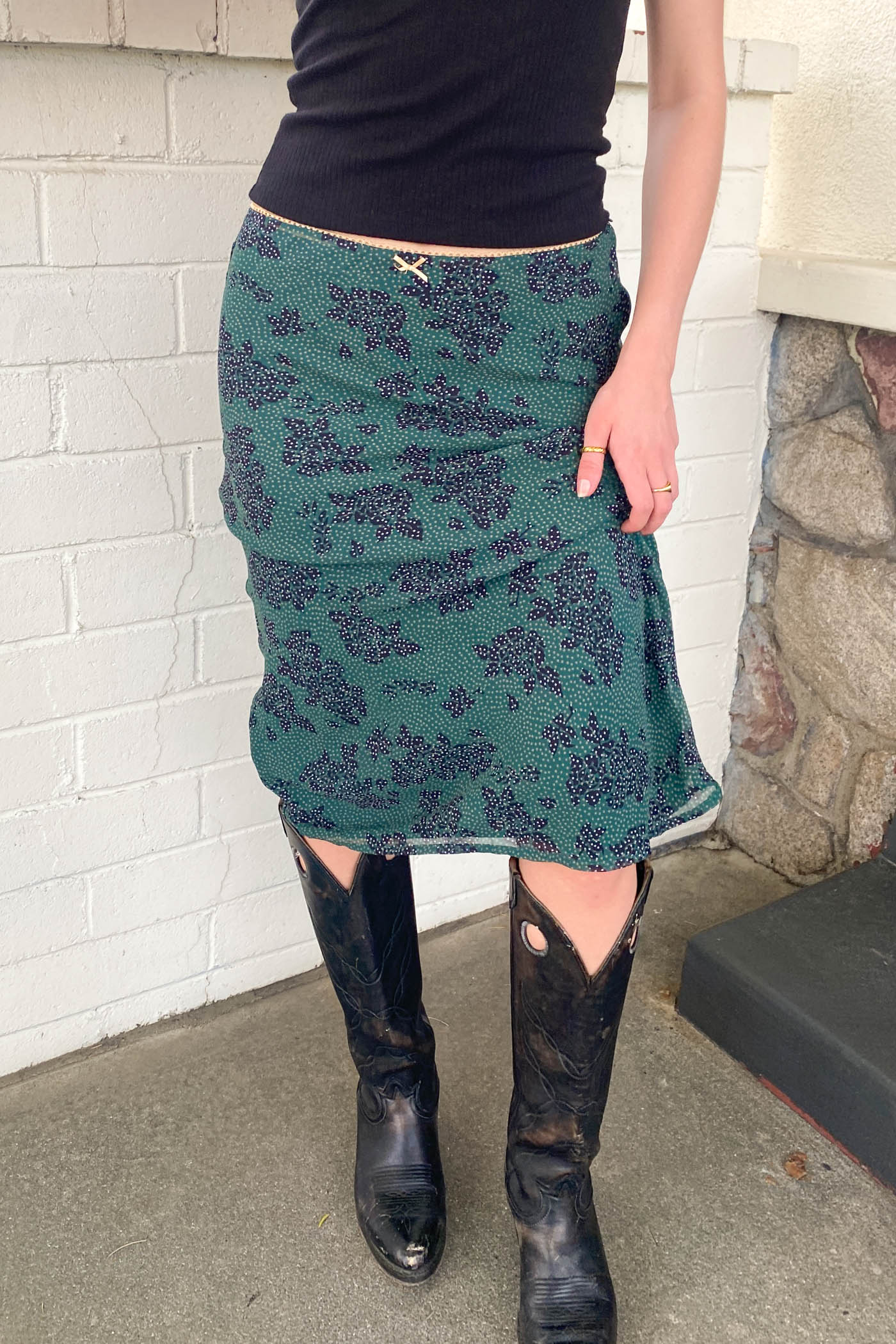 Person wearing a green floral skirt and black boots against a white brick wall.