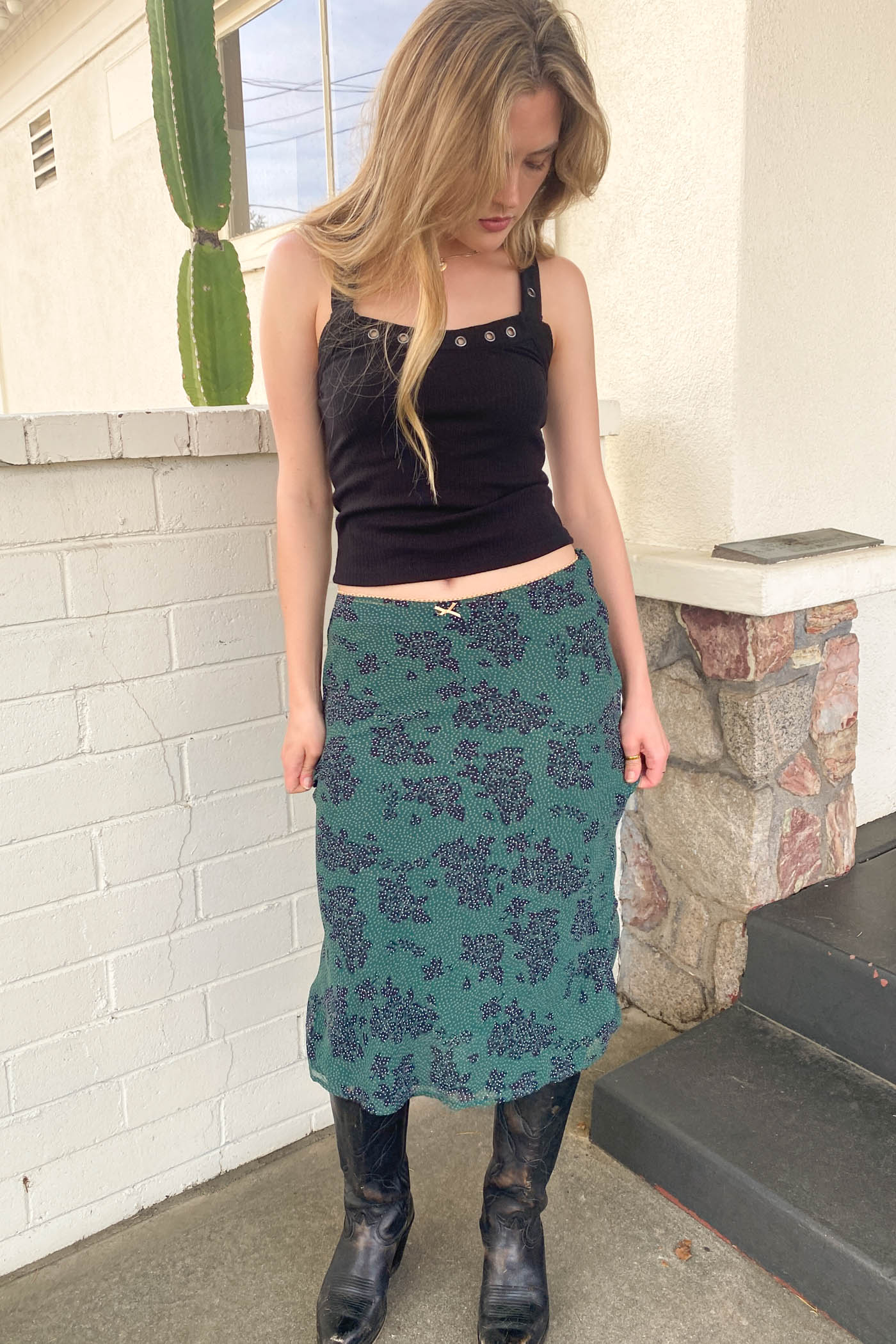 Woman wearing a black top and teal patterned skirt standing on steps with a cactus in the background.
