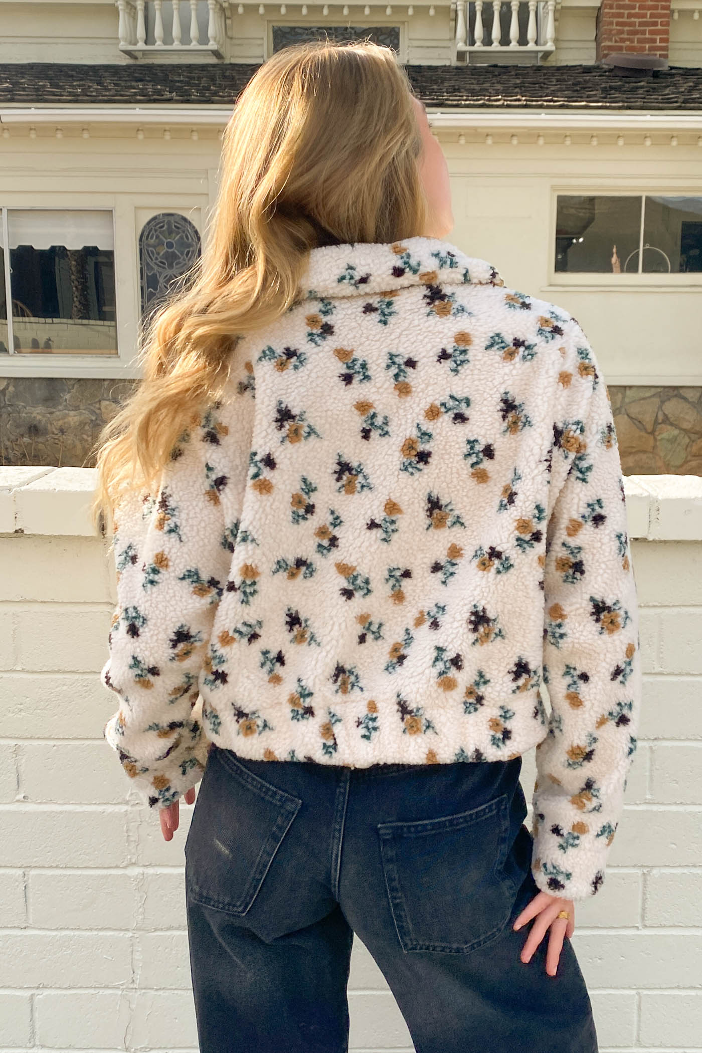 Person wearing a floral shirt and jeans standing in front of a white building.