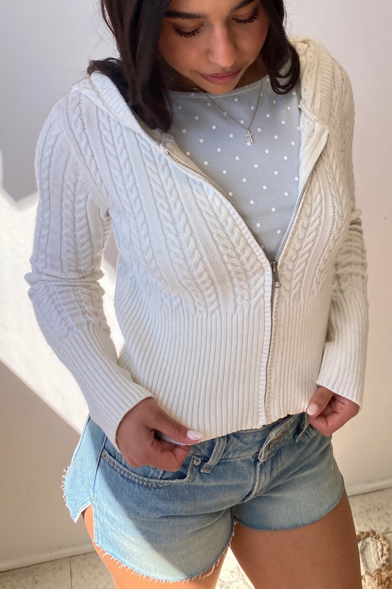 Person wearing a white cable knit cardigan over a light blue polka dot shirt with denim shorts.