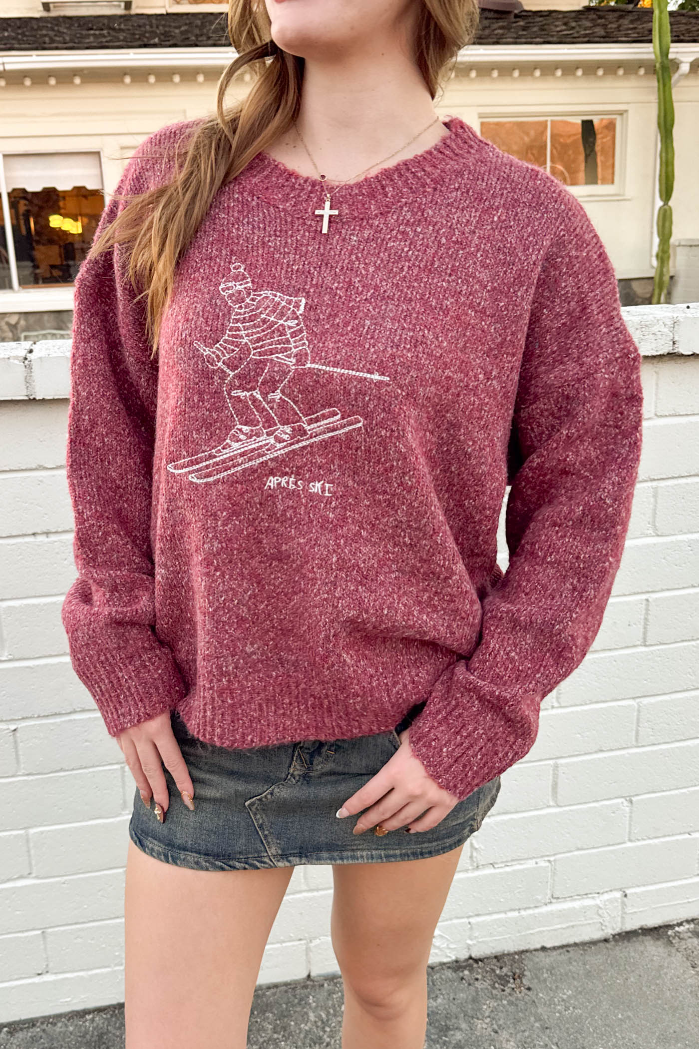 Person wearing a maroon sweater with a graphic design, standing against a white brick wall.