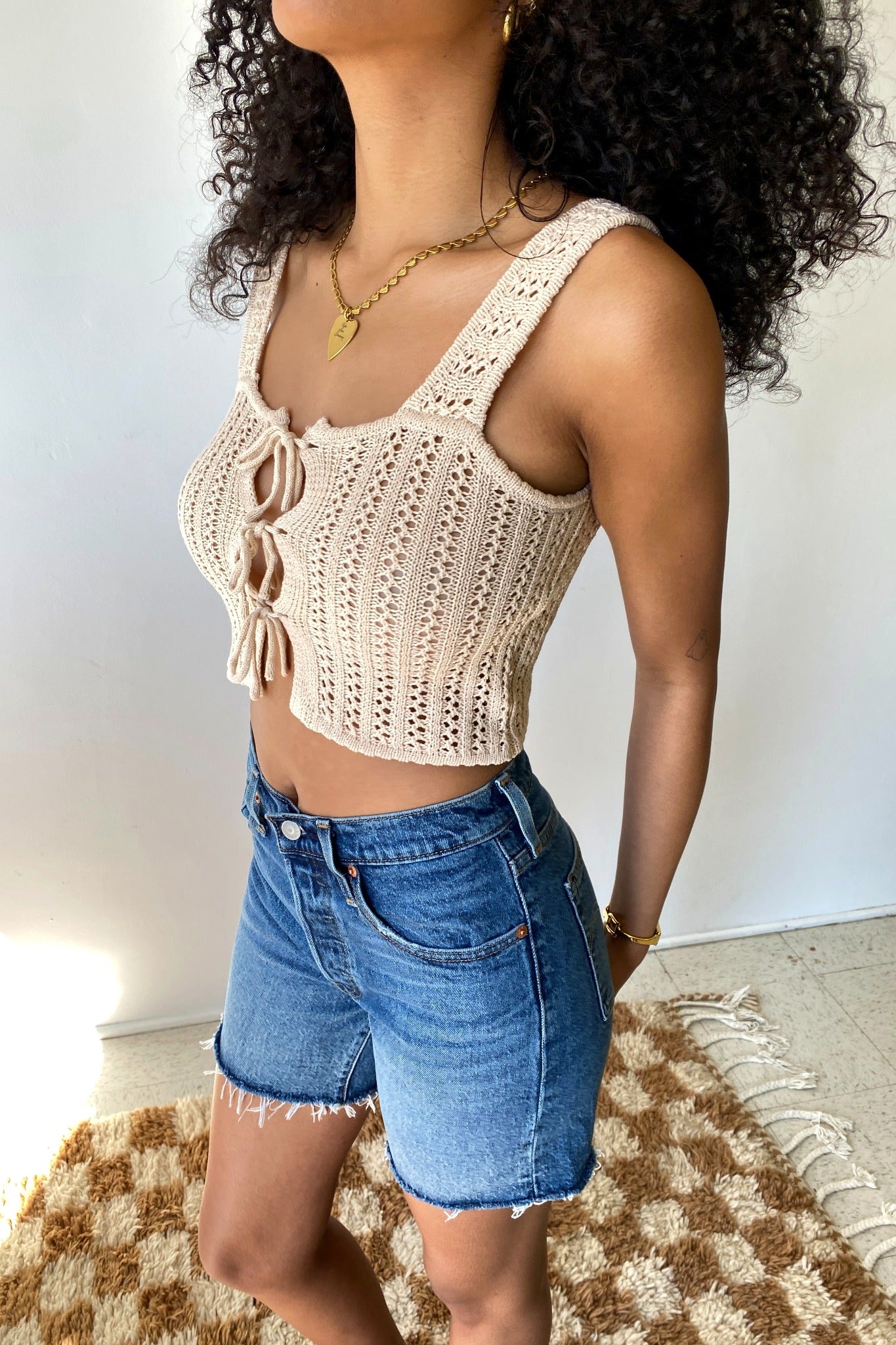 Woman wearing a beige crochet tank top and blue denim shorts against a white wall.