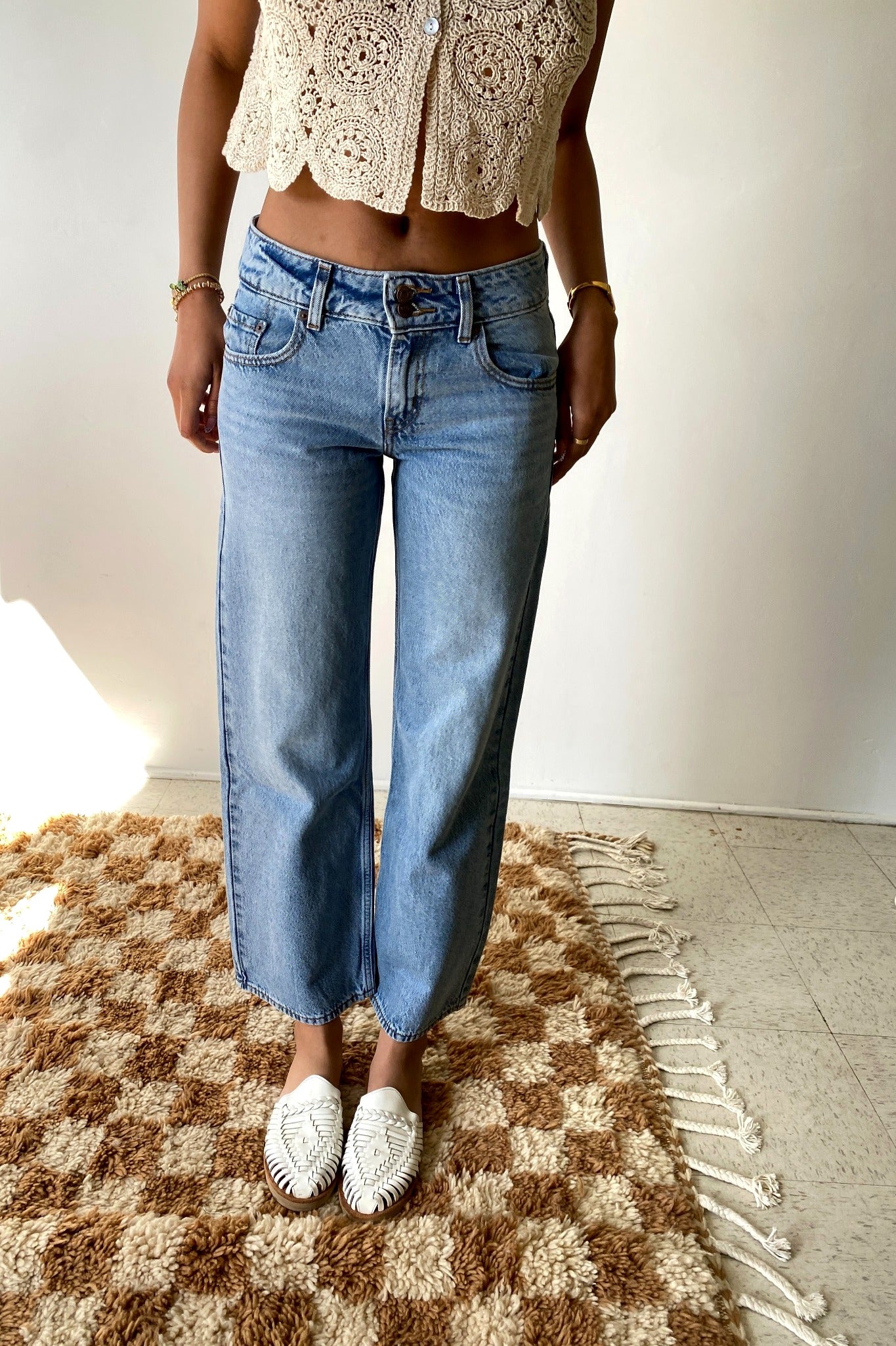 Person wearing a beige crochet top and blue jeans standing on a checkered rug.