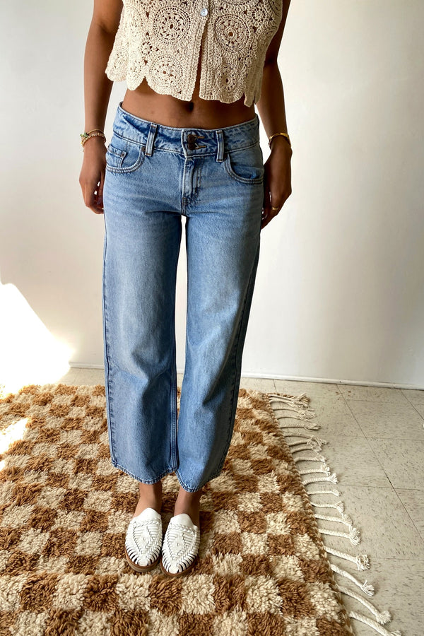 Person wearing a beige crochet top and blue jeans standing on a checkered rug.