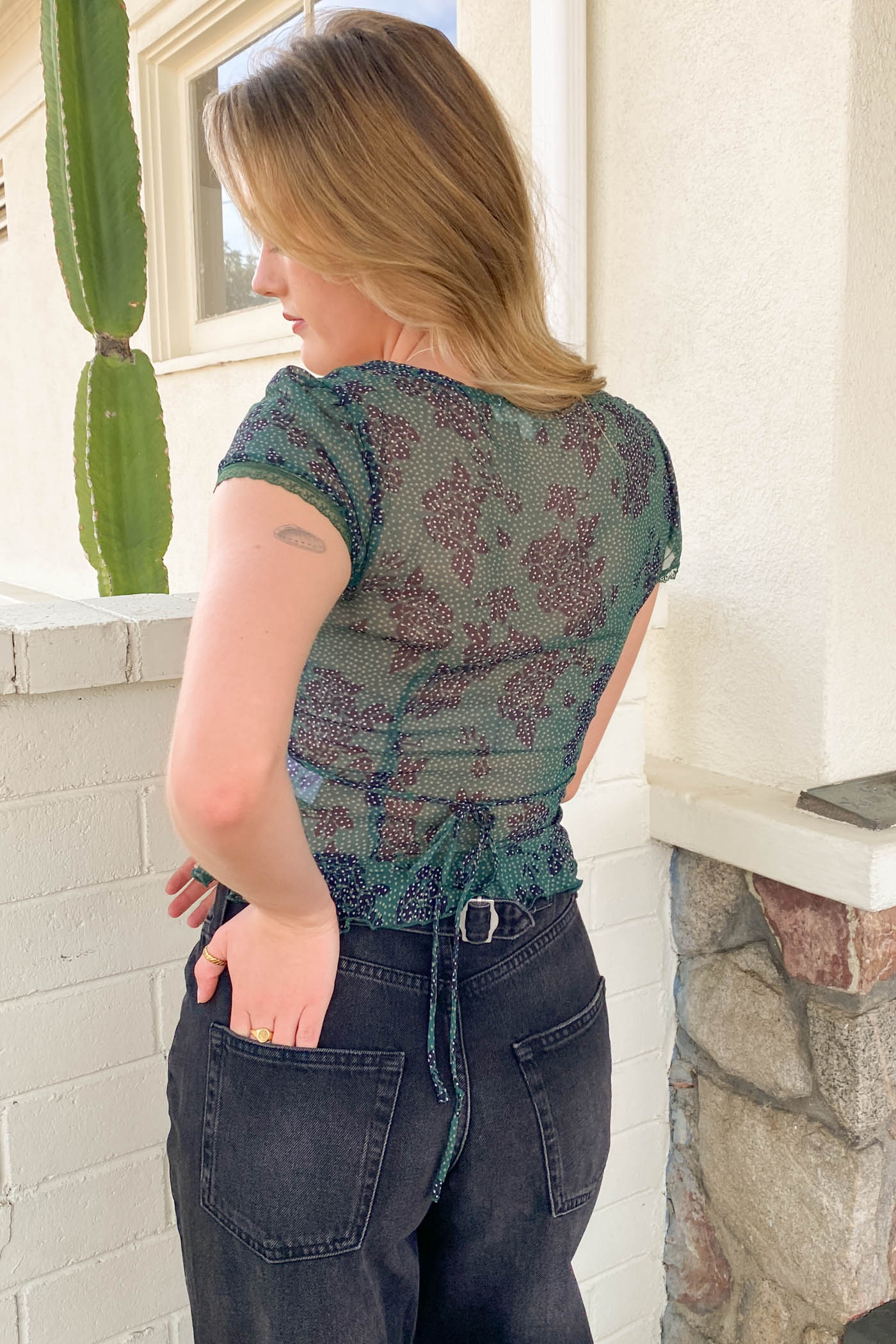 Woman wearing a patterned top and jeans standing in front of a white wall with a cactus plant.