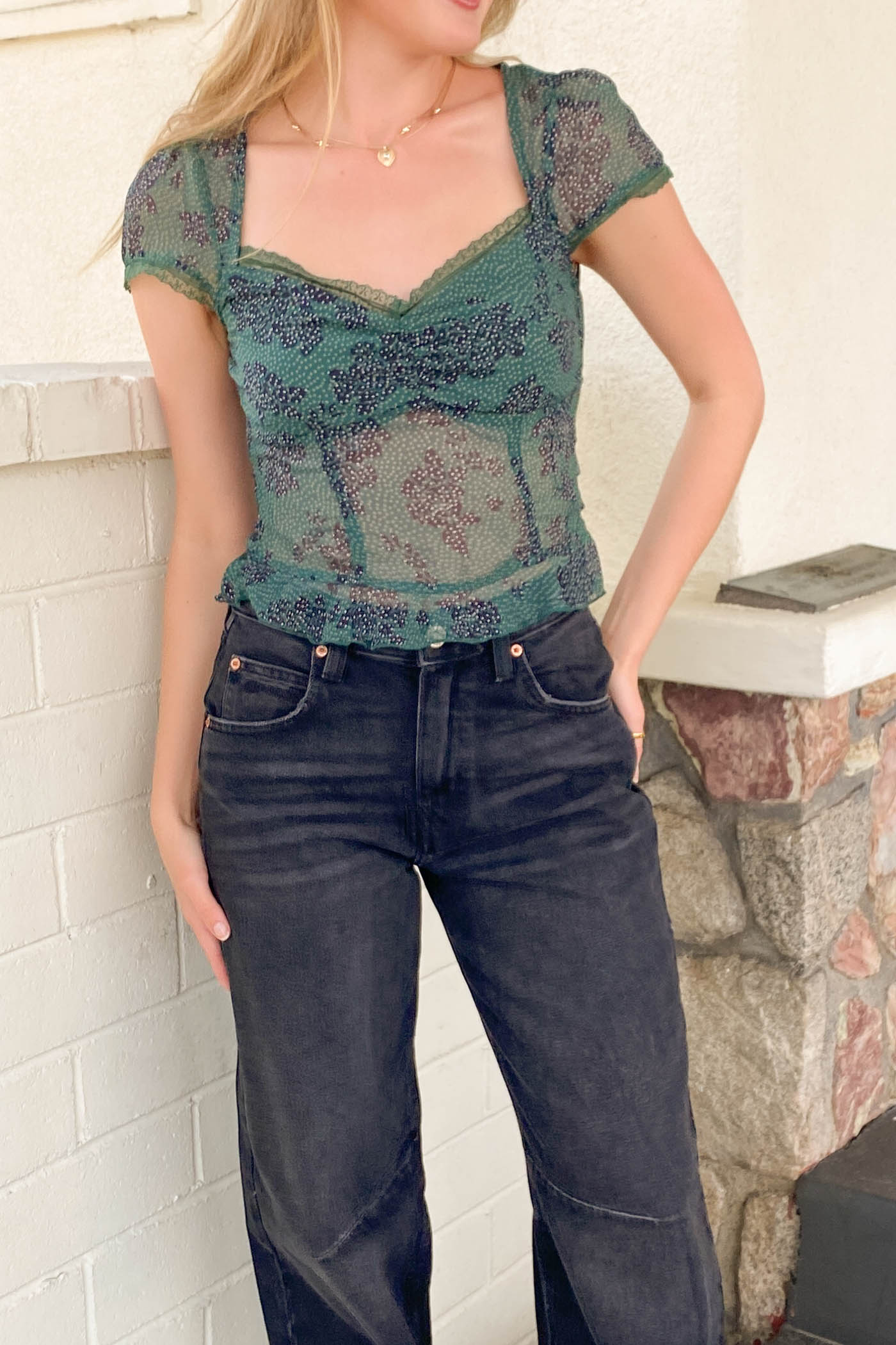 Person wearing a green patterned top and dark jeans against a white brick wall.