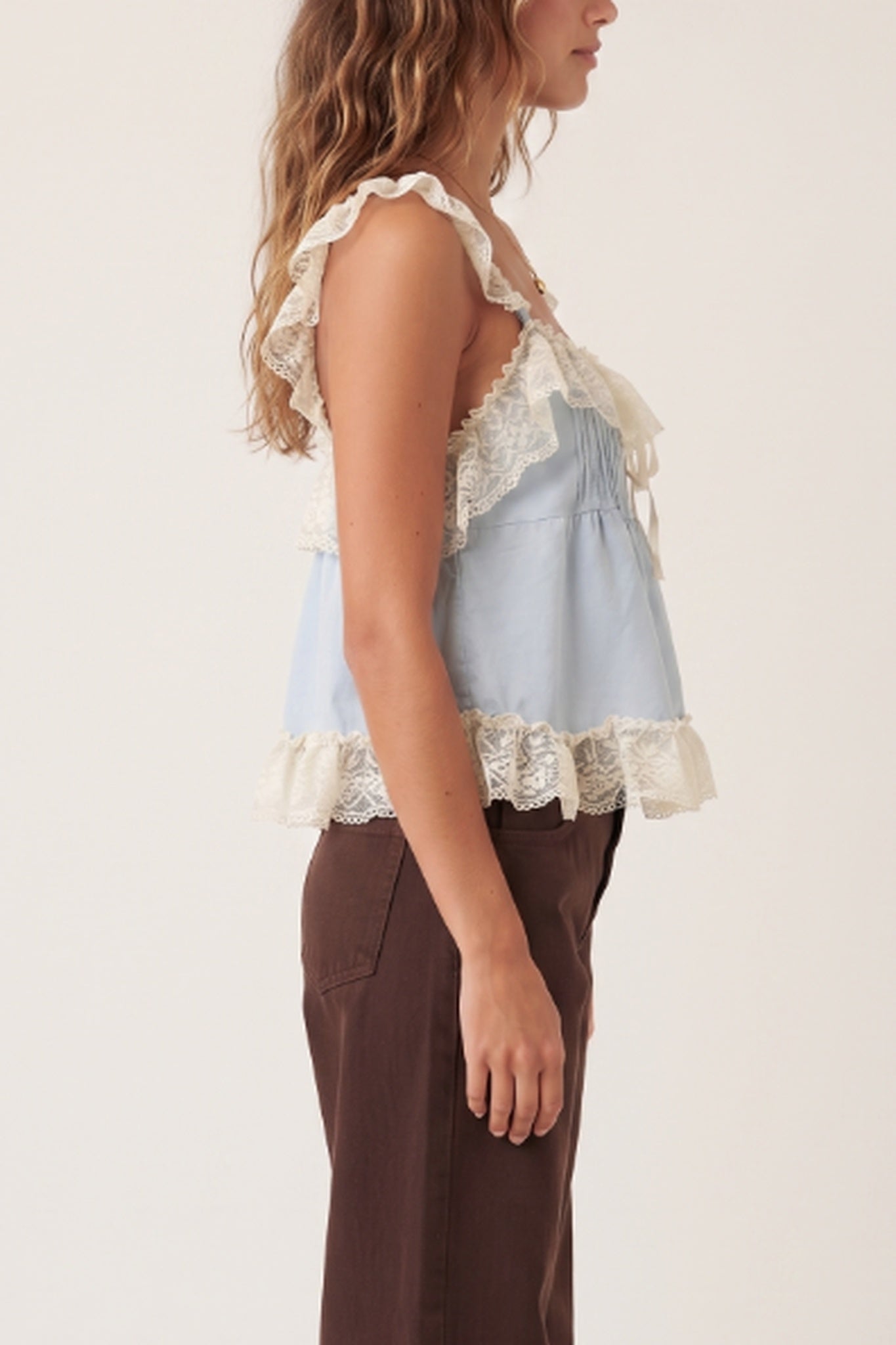 A woman shown in side profile wearing a light blue sleeveless top with cream lace ruffles and dark brown pants, against a plain light background.