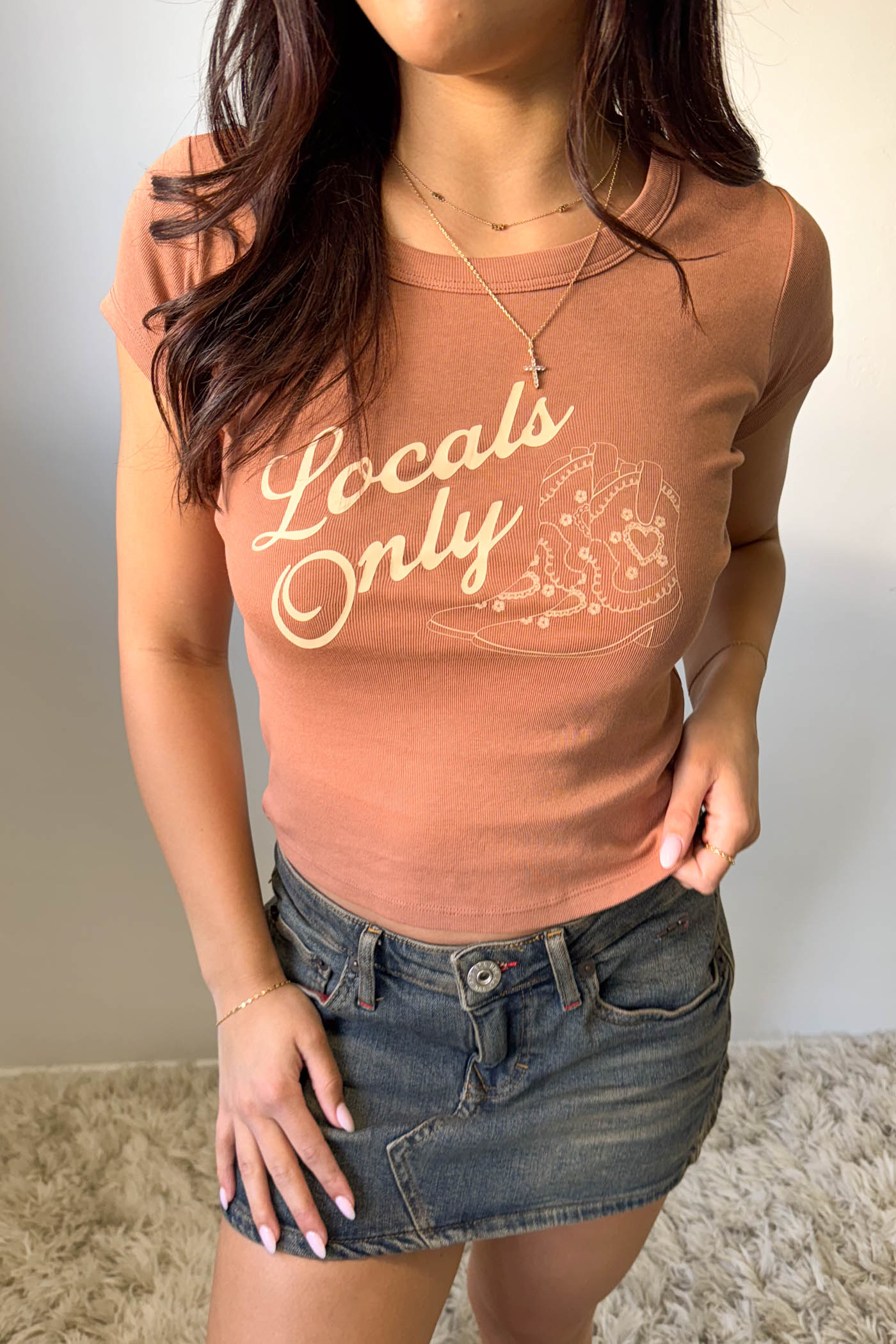 Person wearing a brown 'Locals Only' t-shirt and denim skirt against a neutral background