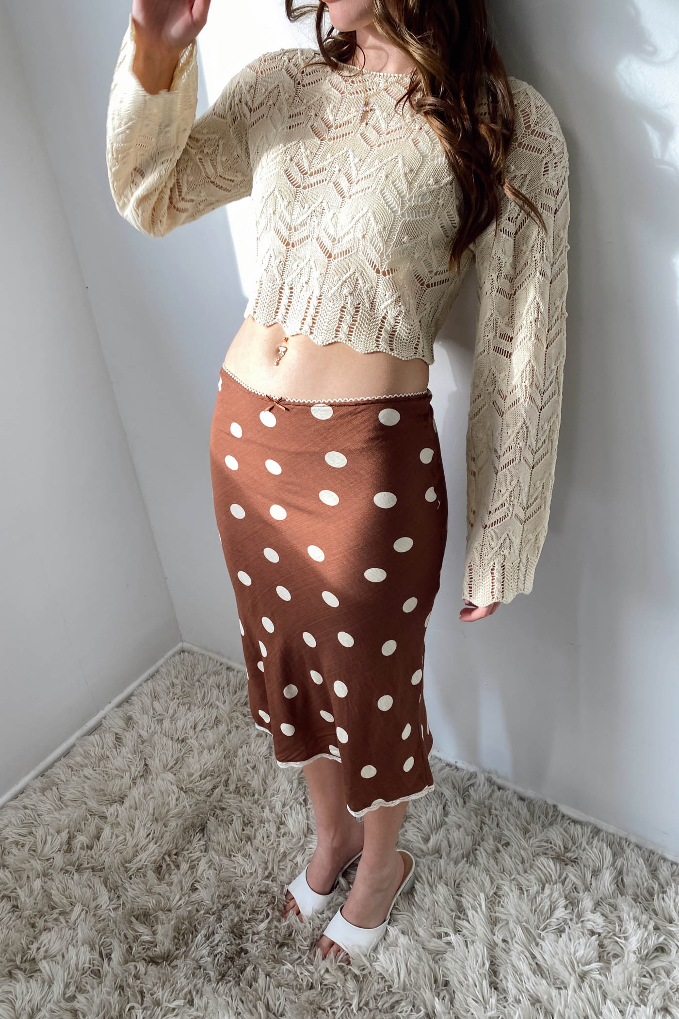 Person wearing a beige lace top and brown polka dot skirt against a white wall.