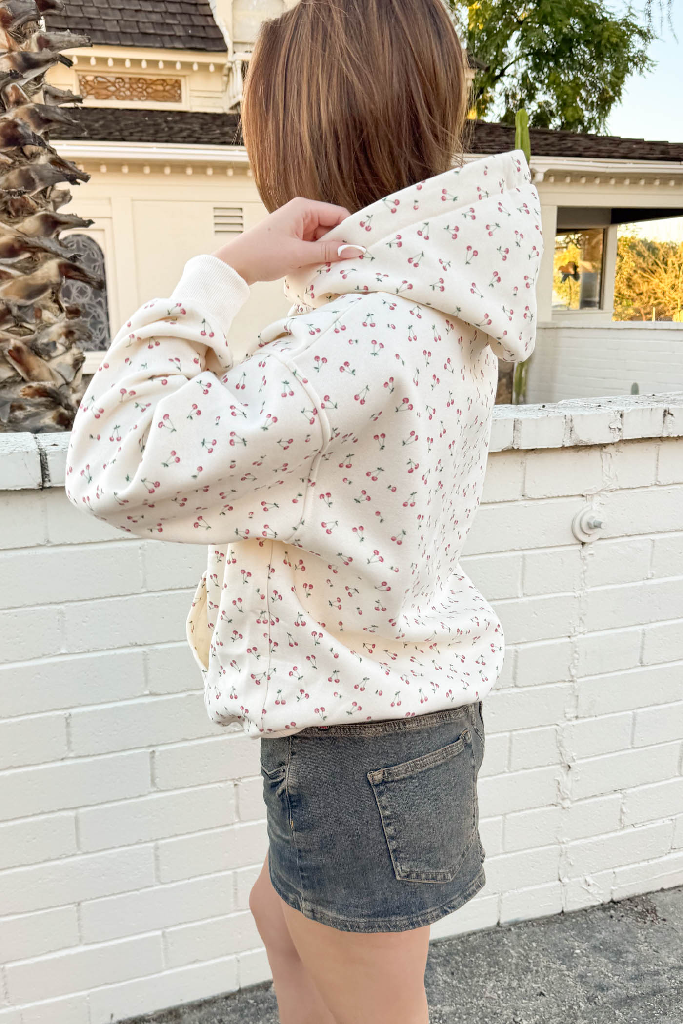 Person wearing a floral hoodie and denim shorts standing in front of a white brick wall.