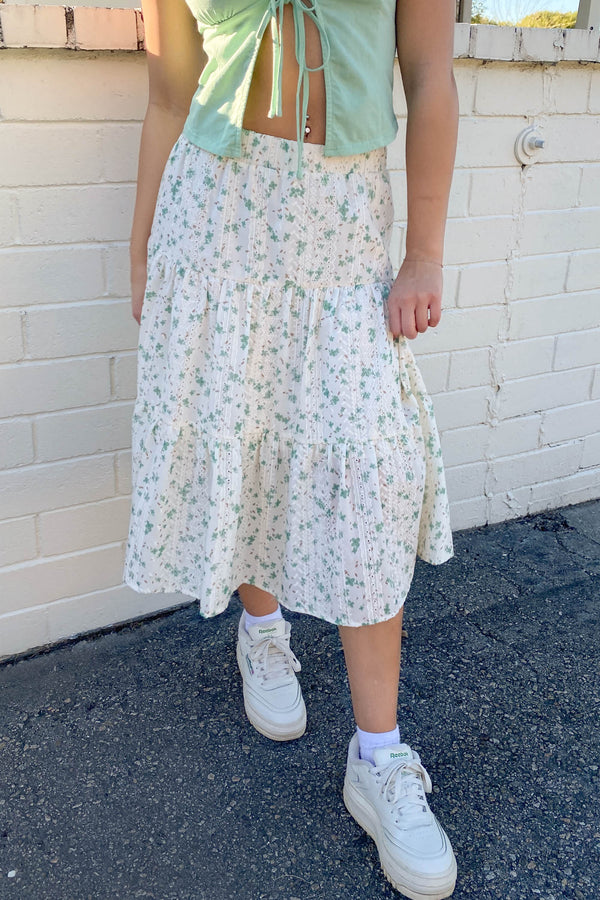 Person wearing a light green top and floral skirt with white sneakers against a white brick wall.