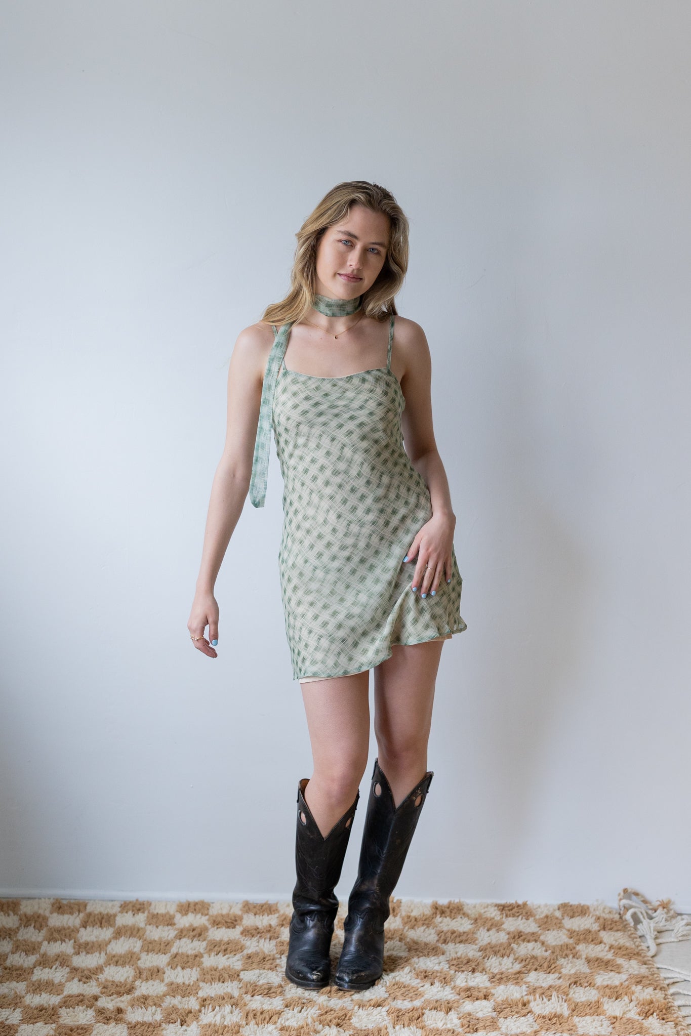 Woman wearing a green patterned dress and black boots standing against a plain white wall.